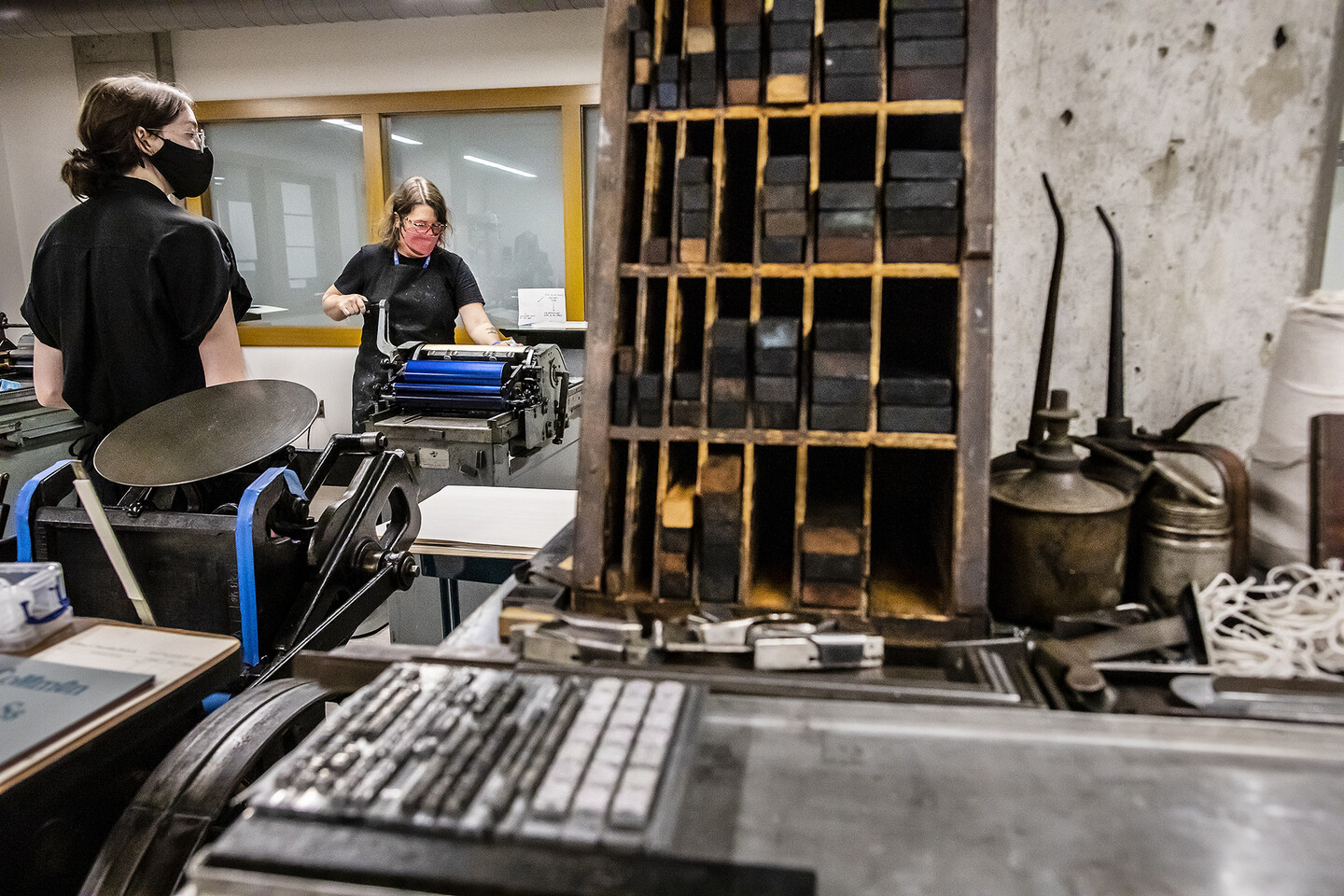 two people working the hand-operated press in the background with wood and metal letters in a shelf in the foreground