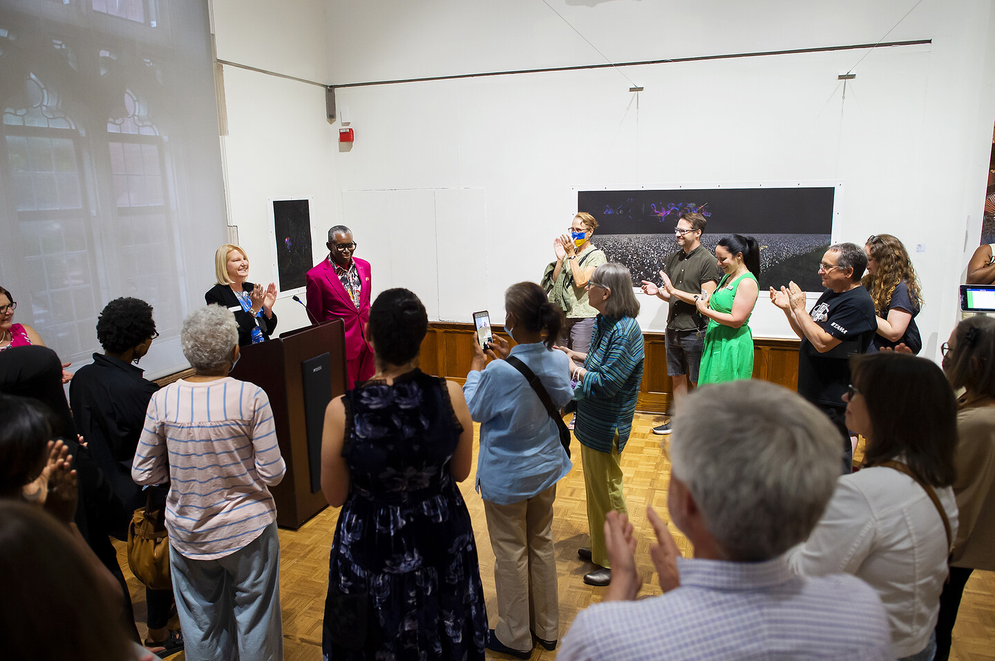 curator Lynn Marsden-Atlass and artist John Dowell standing at podium in gallery with several people standing in front of them clapping