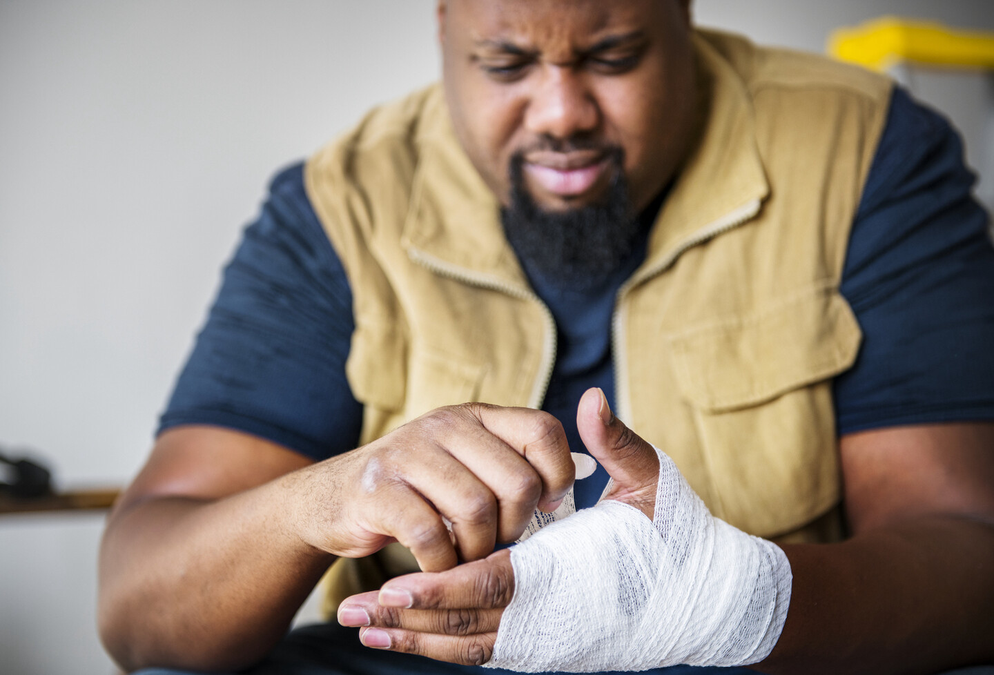 Person with a goatee and moustache wearing a tee-shirt and vest, the hand wrapped up in gauze as if it was injured. The person has a pained expression.