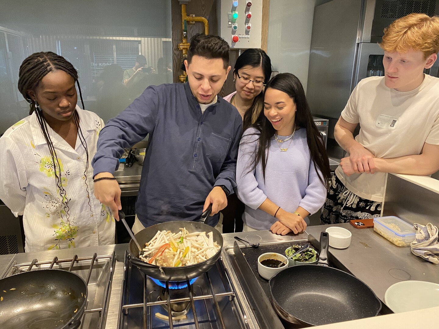 Four students watch a a cooking demonstration as a man stirs mushrooms and peppers in a wok on a gas stove.