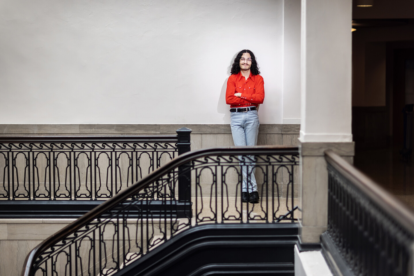 A student in a red long sleeved shirt stands with arms crossed leaning against the wall atop a staircase.