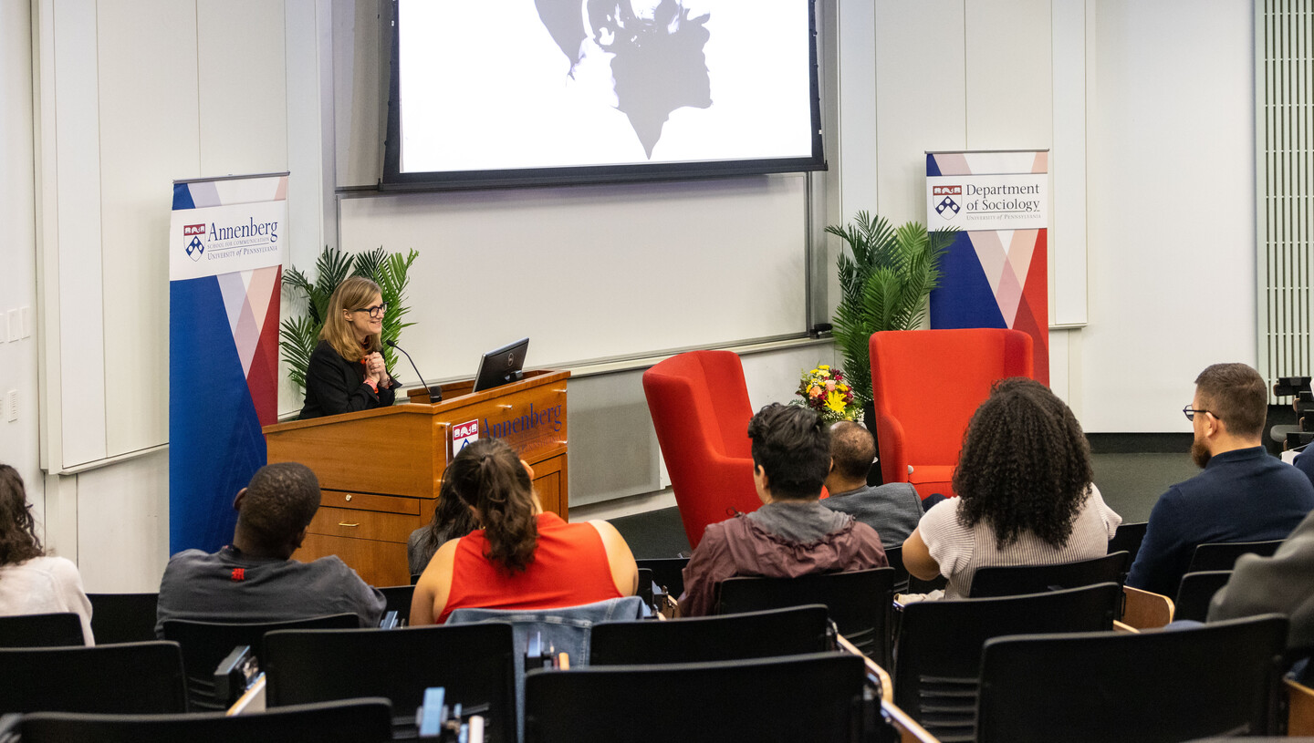 Penn president Liz Magill standing at a lecturn in a small auditorium. Visible are the backs of heads sitting in the rows near her, as well as signs for the Annenberg School for Communication and Department of Sociology.