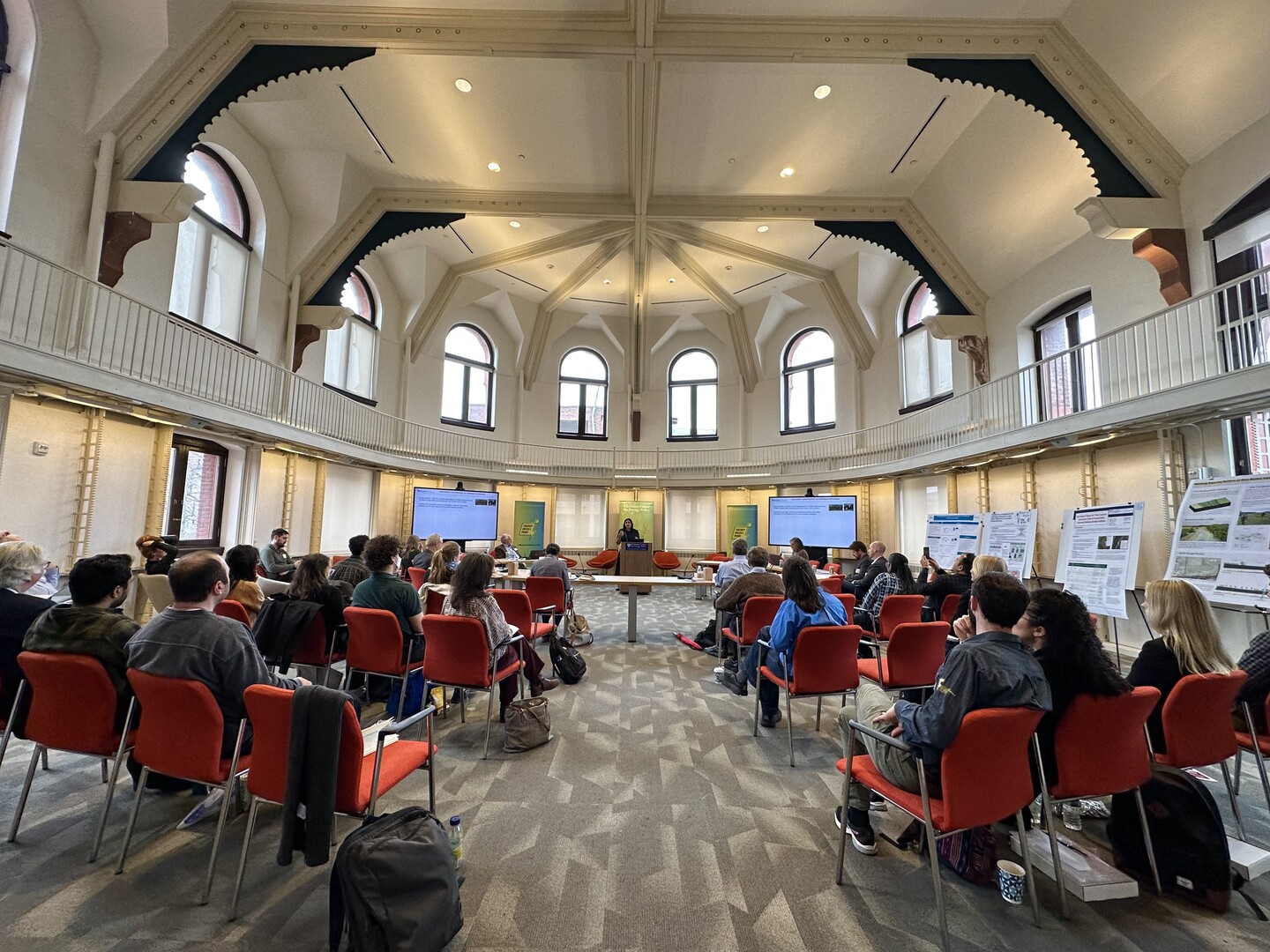 Person speaks at a podium in an airy conference room as a couple dozen people listen
