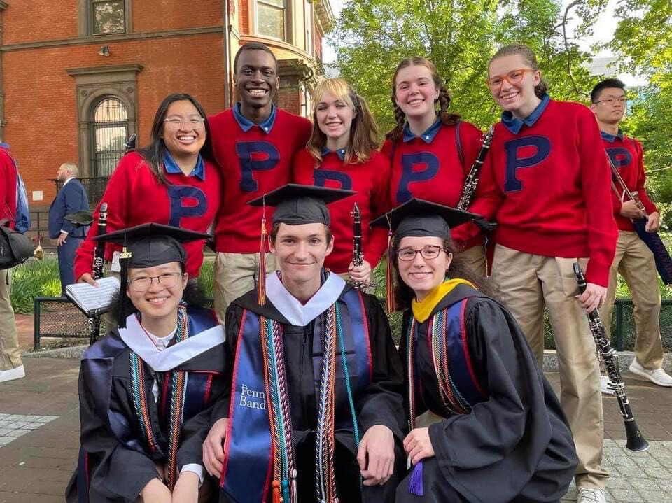 Five people wearing red sweaters with an applique "P" holding clarinets. In front of them, three graduating bandmates sport gowns and regalia