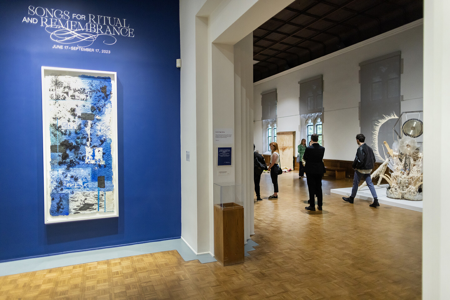 artwork on wall with title Songs for Ritual and Remembrance above and people in gallery behind 