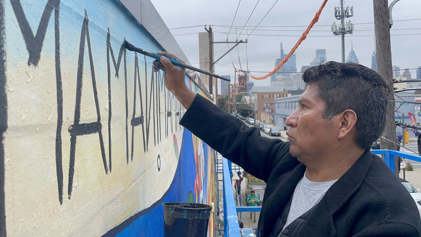 Mamani Mamani signs his name to the work in black paint. The city of Philadelphia is in view in the background
