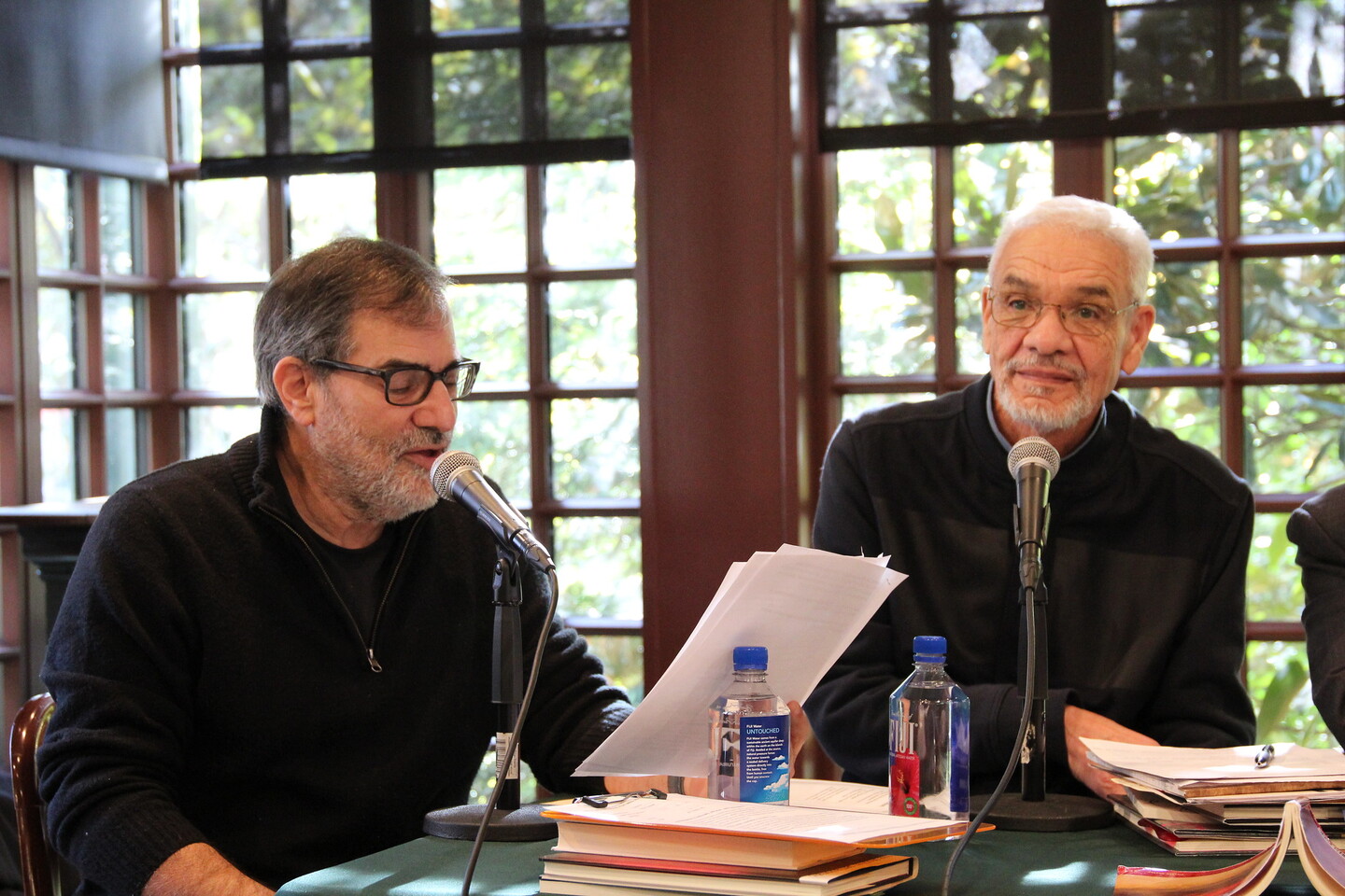 Al Filreis looking at sheets of paper while speaking into a microphone seated at a table with windows behind