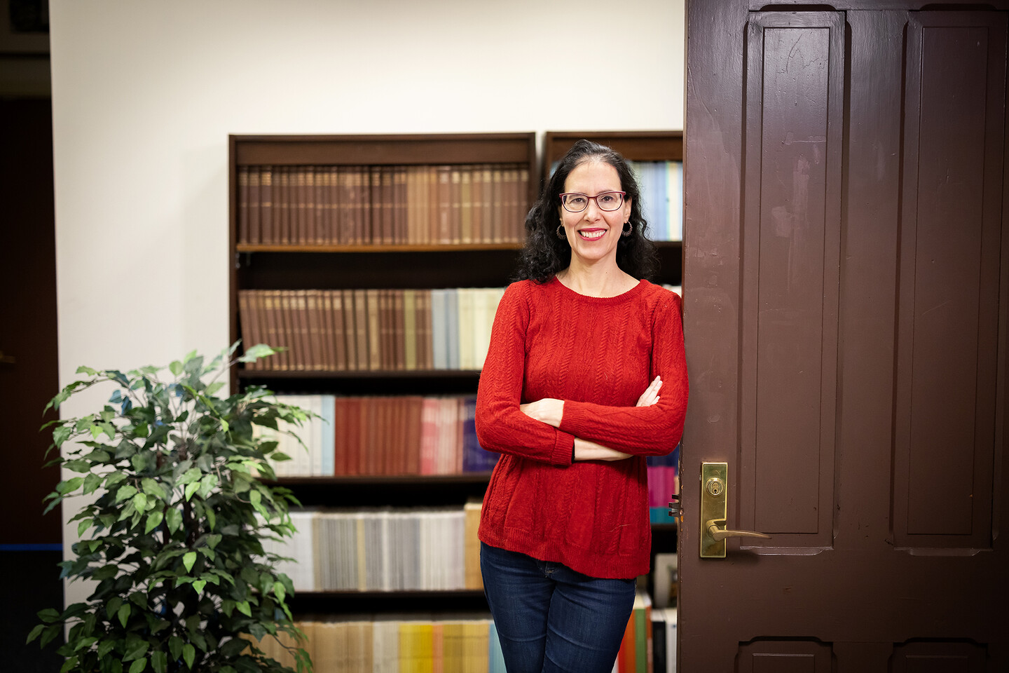Historian Marcy Norton crosses arms in front of chest and leans against a dark wood door with bookshelves behind her.