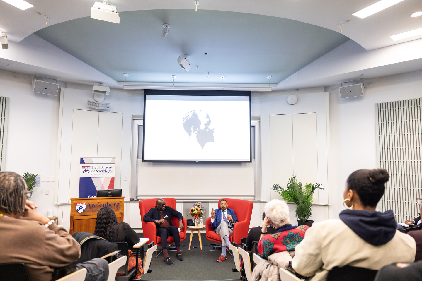 Zuberi and Morris sit on red chairs at the front of a large lecture hall. The podium reads "Annenberg" and there is a sign saying "Department of Sociology." 