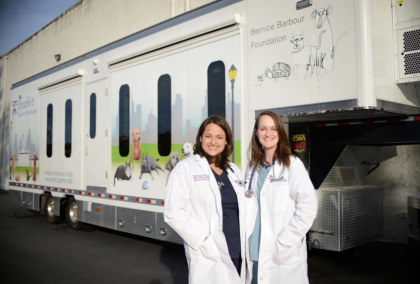 Two doctors in white coats stand in front of the mobile shelter unit