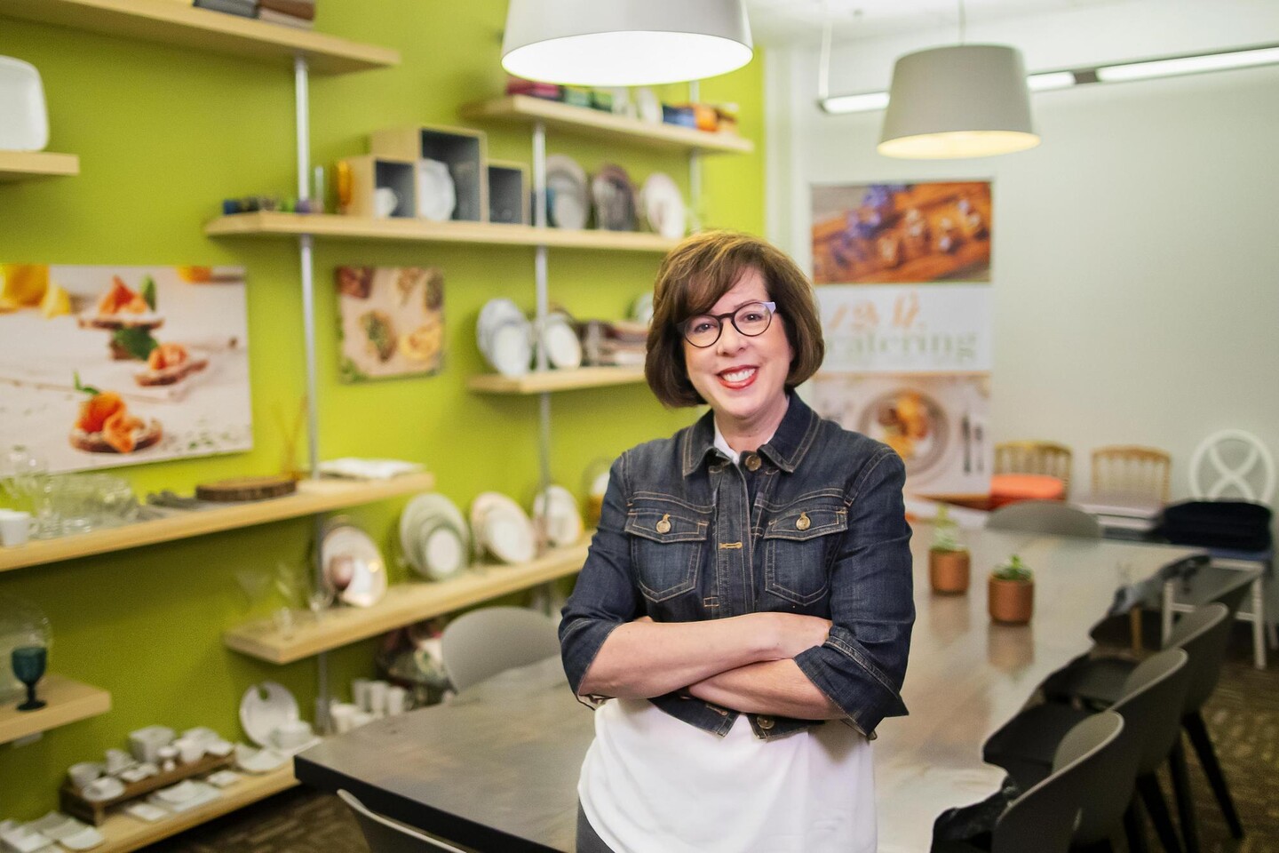 Michele Leff, founder of 12th Street Catering, a West Philadelphia-based catering company, standing in a brightly-colored kitchen with dishware and dining utensils on display.