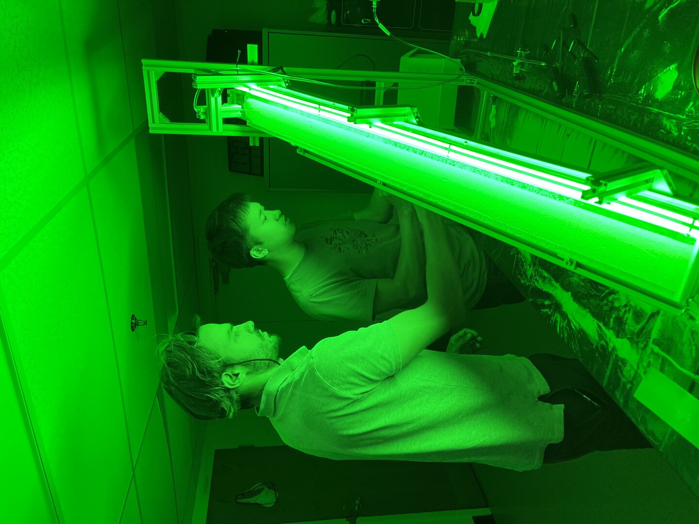 Two researchers stand beside a sloped, sand-filled experimental channel illuminated by green light, monitoring a setup used to study raindrops rolling downhill.