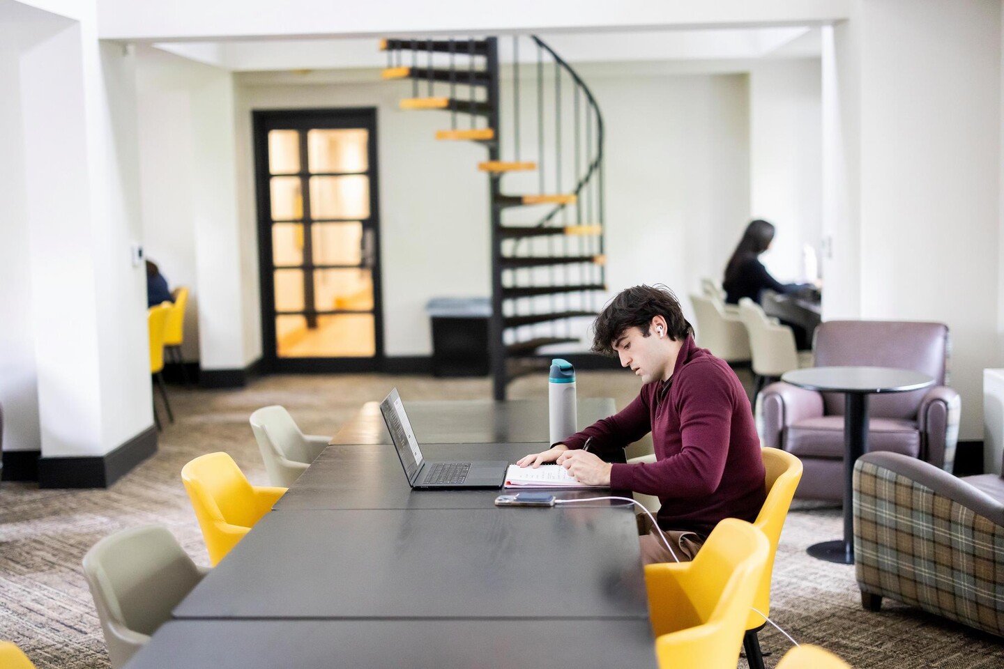Student studying in a lounge with a laptop and notepad.