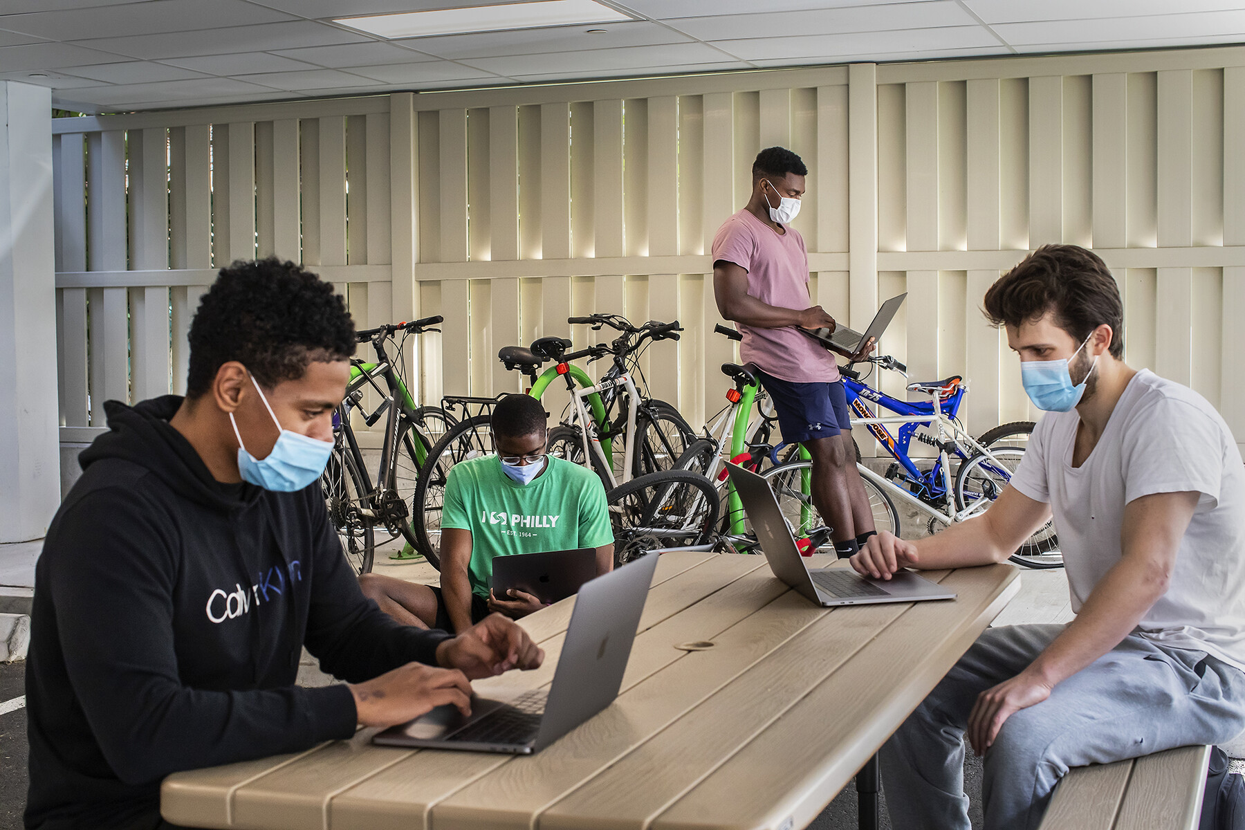Four Penn students in off-campus housing wear face masks and sit at a picnic table looking at laptops.