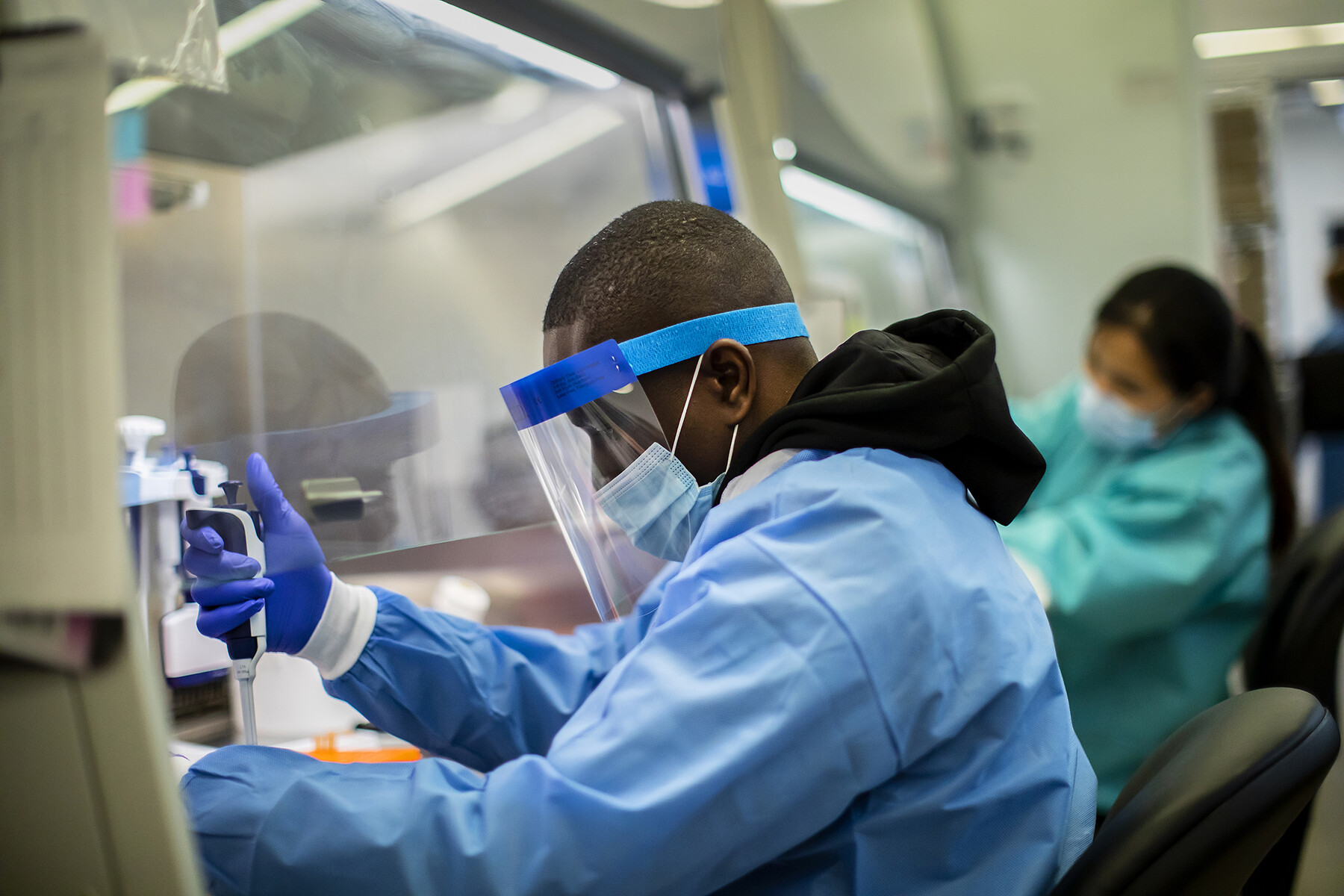 Person seated in a lab wearing full PPE handling a handheld device.