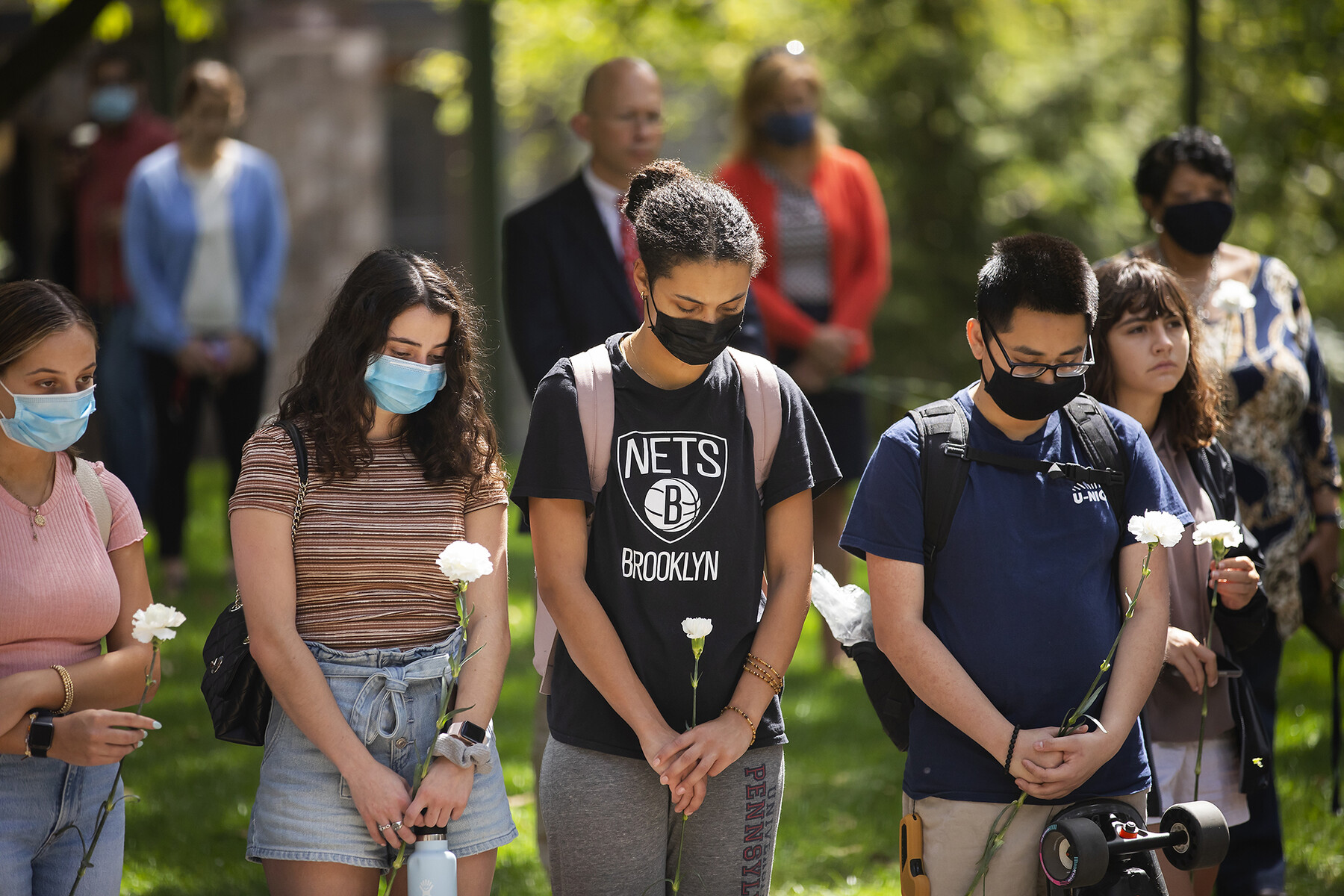 Several. masked students standing on College Green with heads bowed holding white carnations.