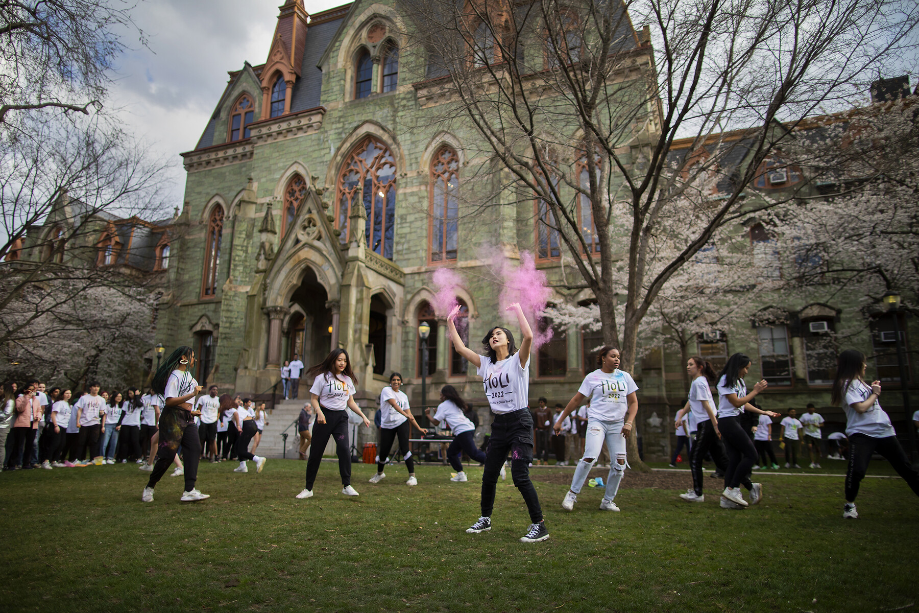 A person wearing a Holi t-shirt throws pink powder into the air surrounded by other students in front of College Hall.
