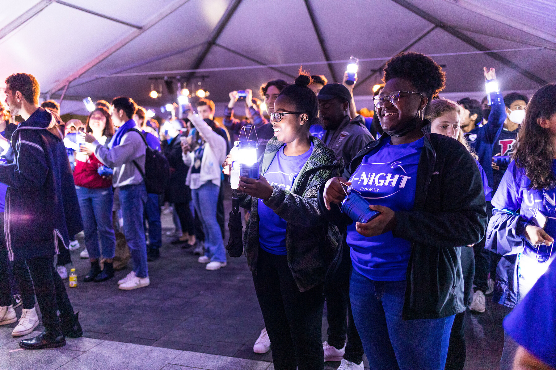 A group of students in a large tent hold lit lanterns in the air.