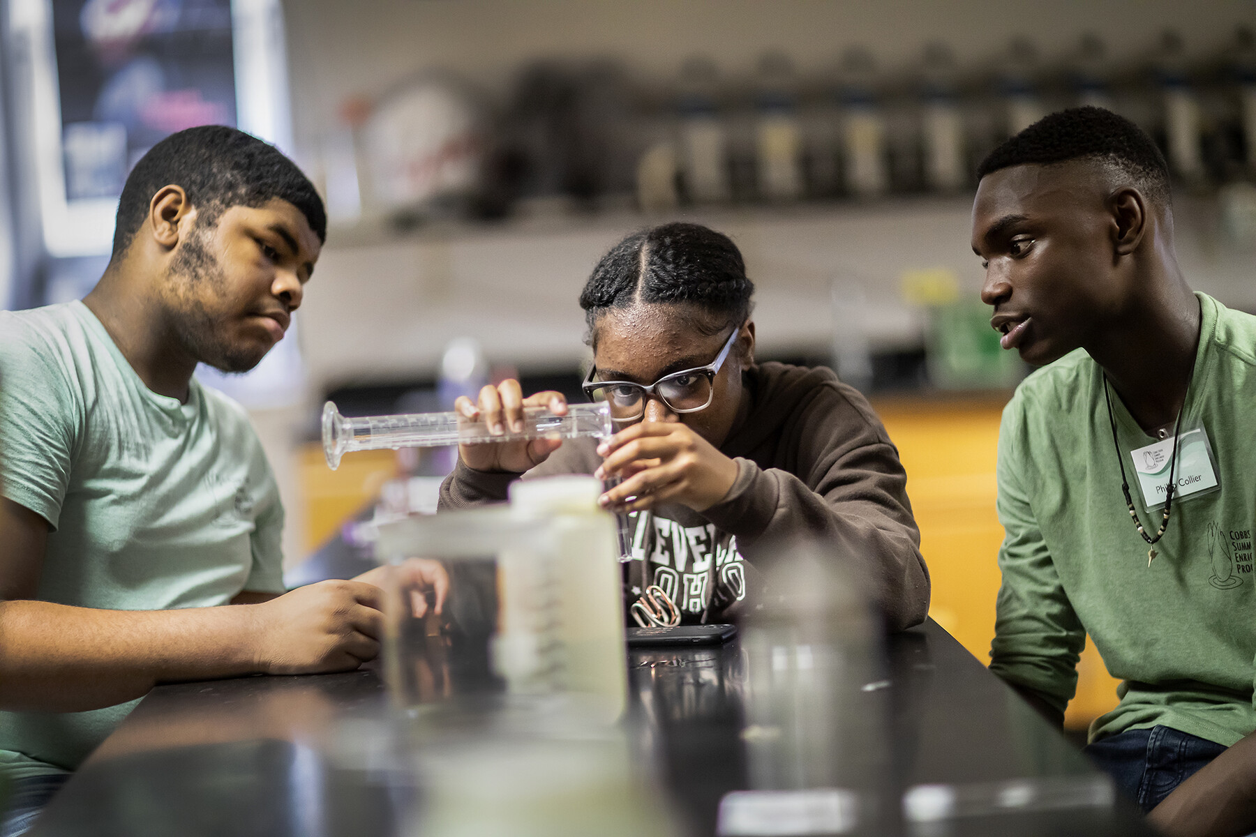 Three students in a lab, one purs liquid from a beaker into a test tube.