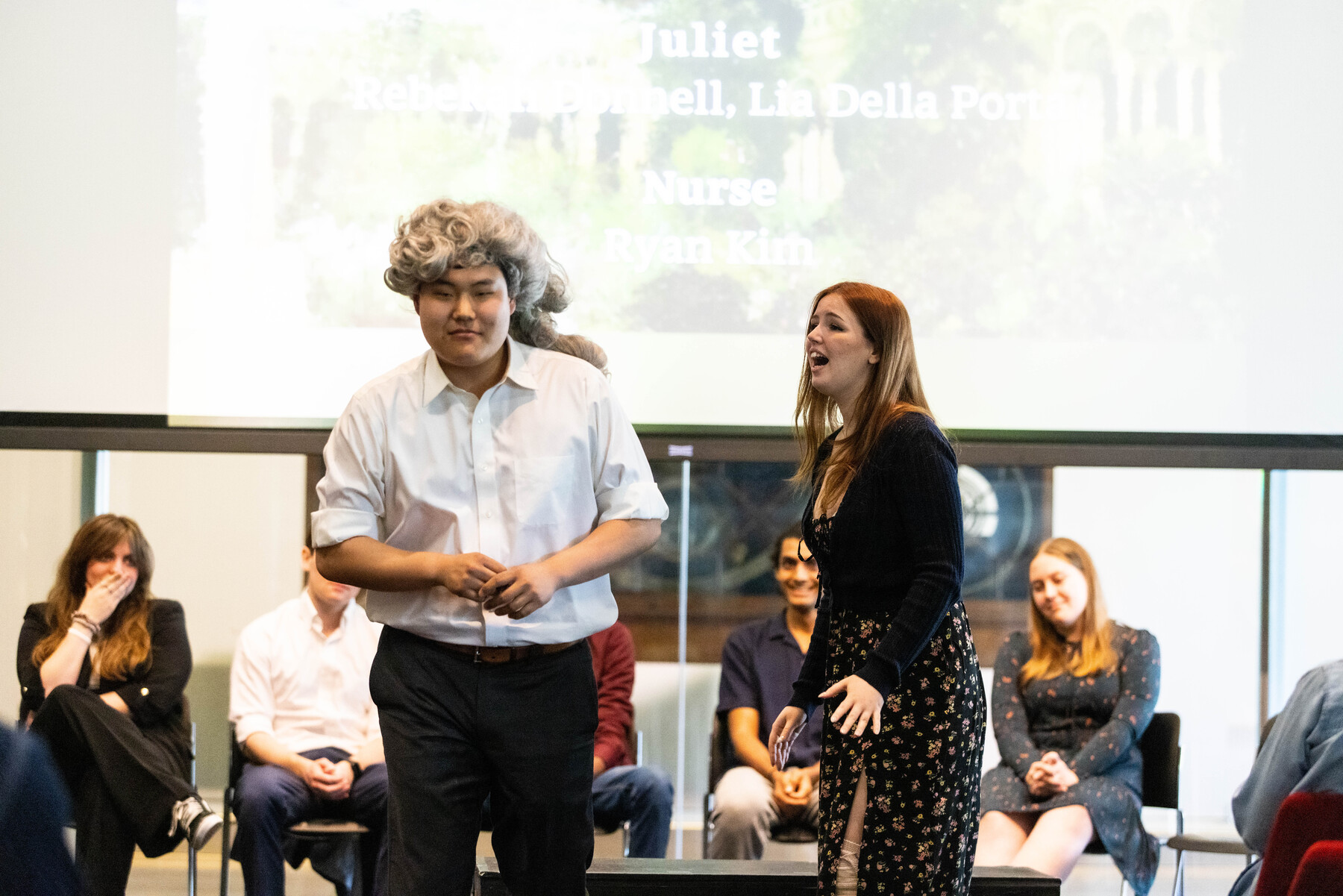 two students in front of an audience, one wearing a wig