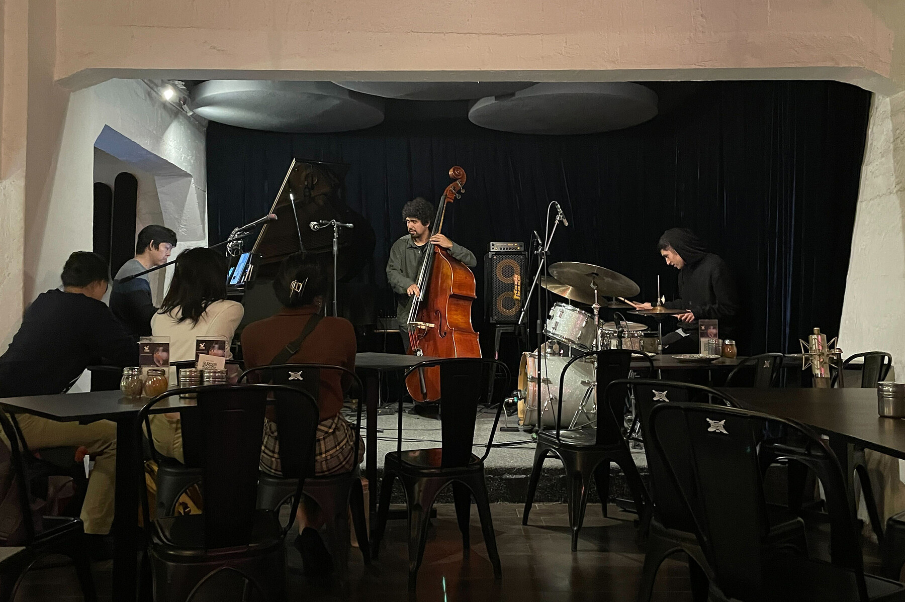 A jazz trio playing instruments as three people in the audience listens.