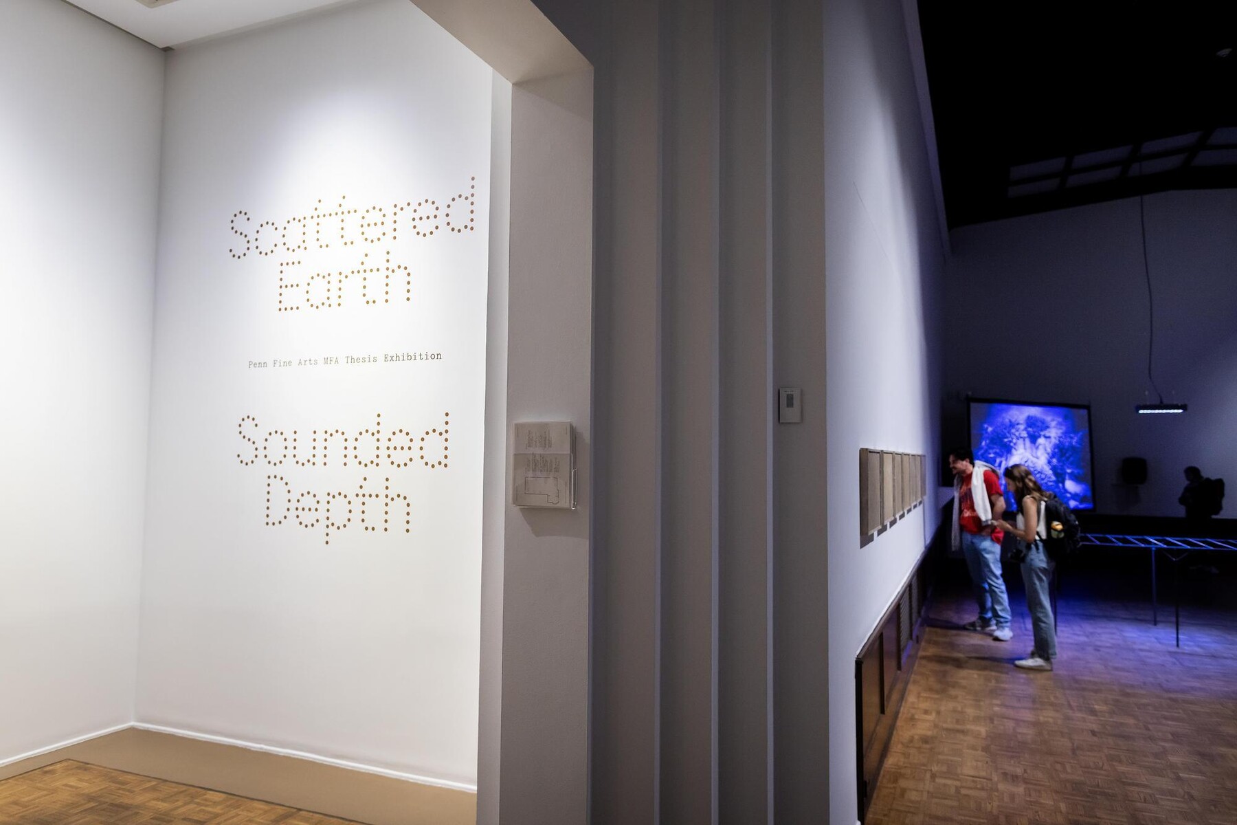 Entrance sign to Scattered Earth Sounded Depth and people looking at artworks on a wall with a video playing on a screen in the back of the gallery 