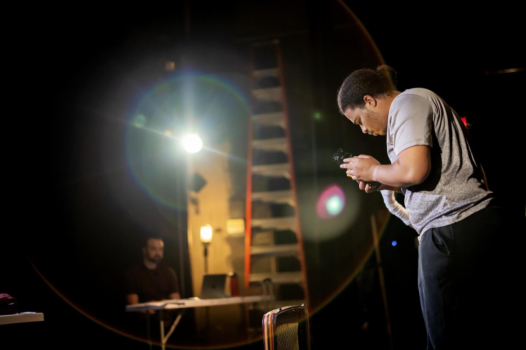Elliot Ross-Dick stares down at a held oil lamp, with Noah Levine and a flaring stage light in the background.