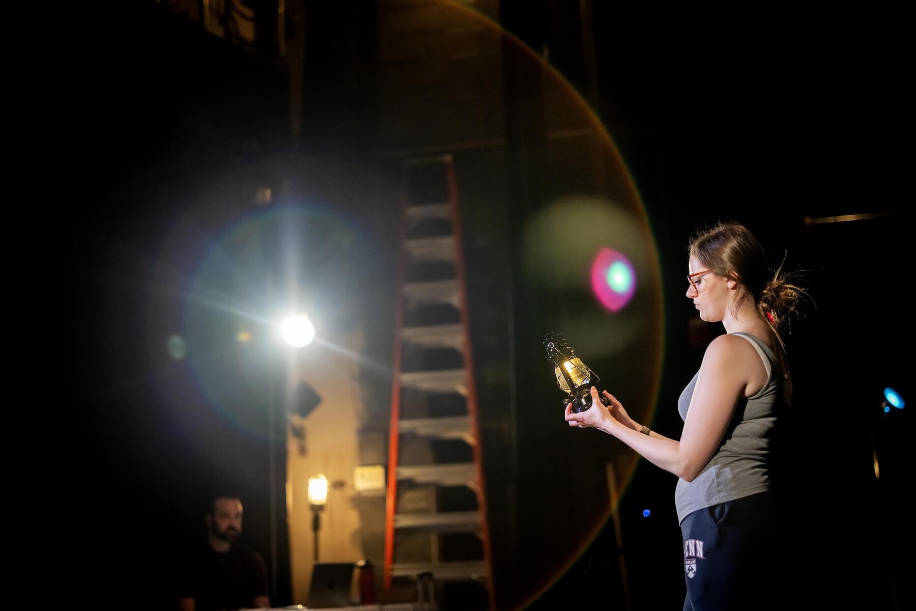 Pandora Schoen stares down at a held oil lamp, with Noah Levine and a flaring stage light in the background.