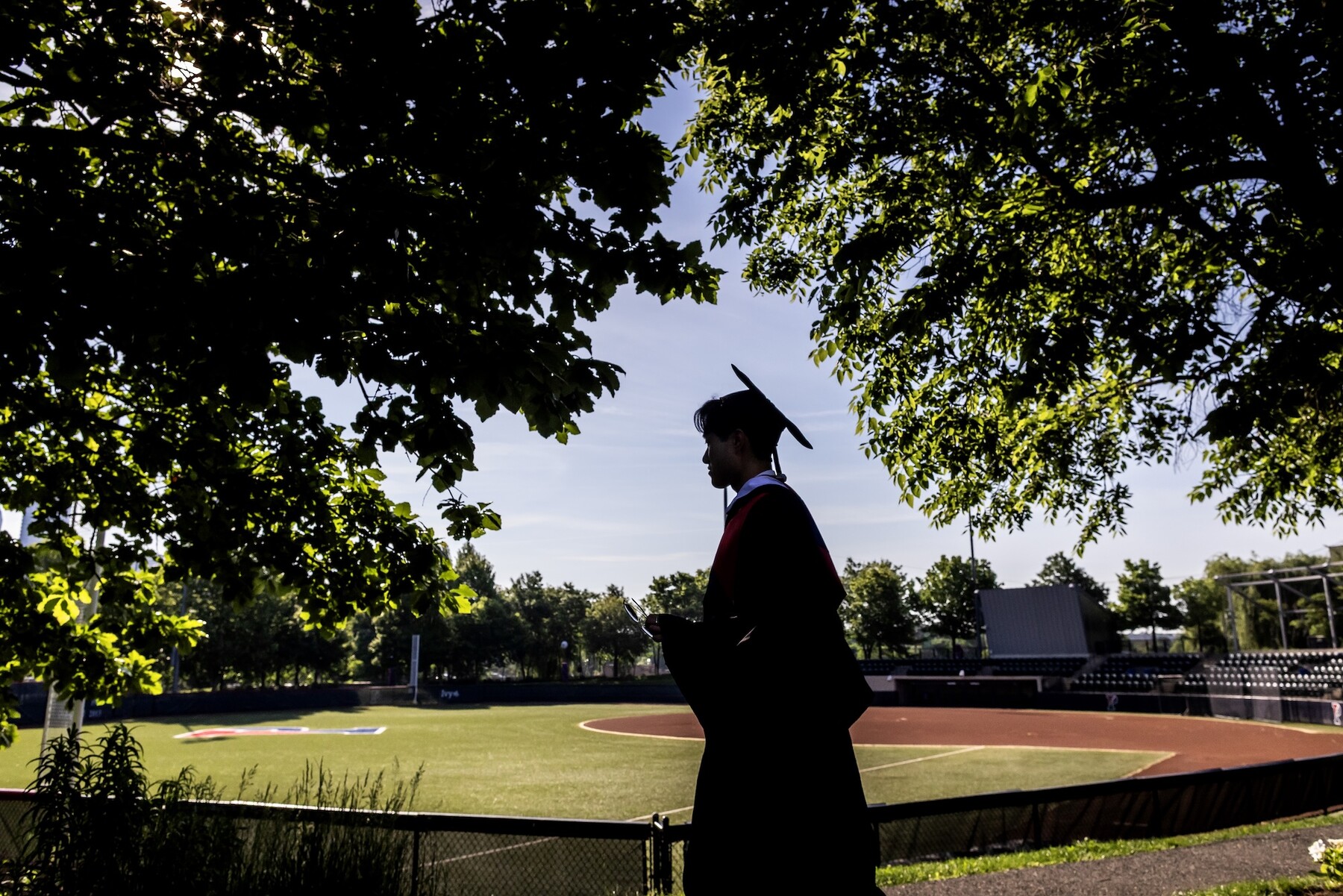 A graduate walking past the softball field in regalia.