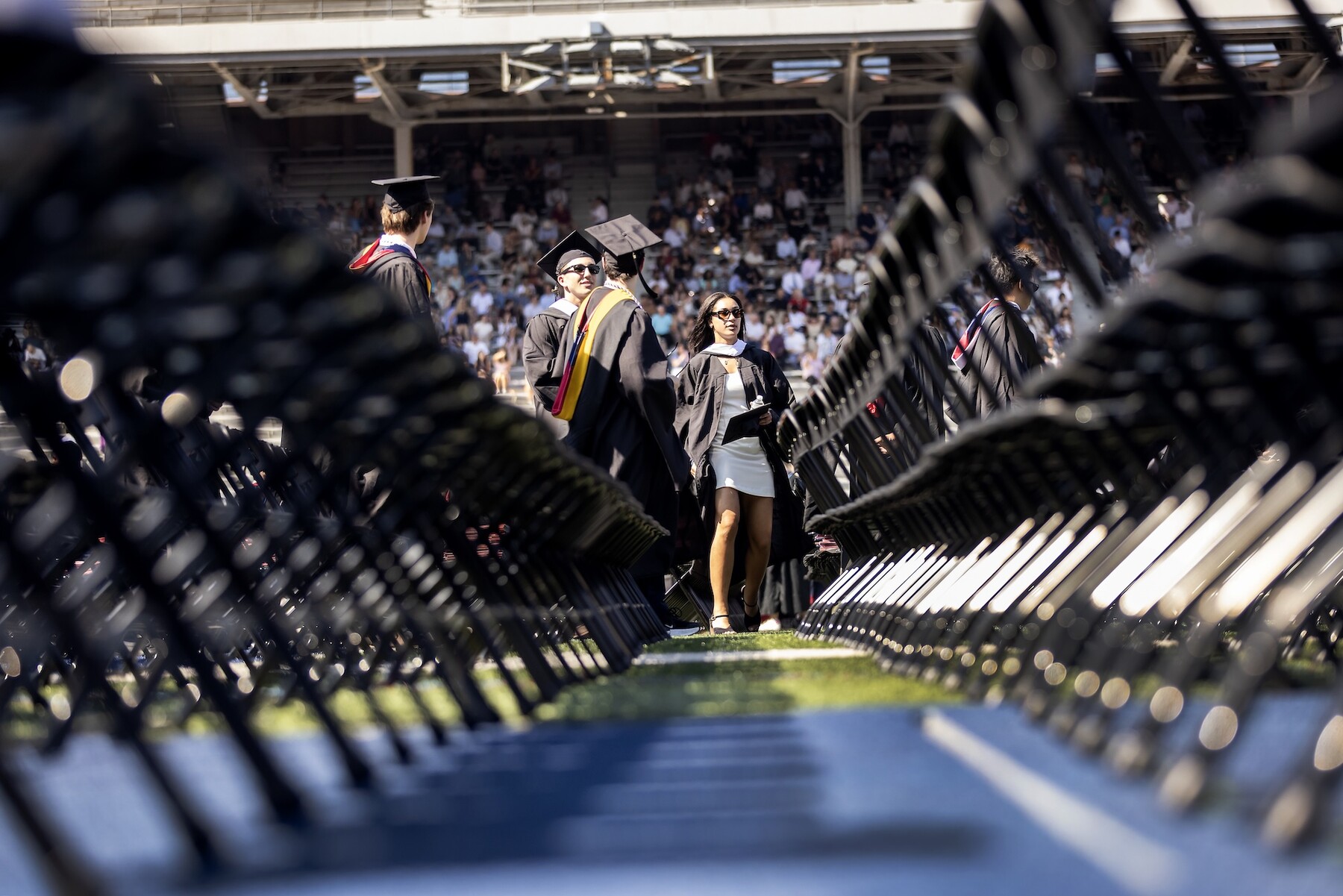 Students walking onto Franklin Field seen through the legs of the folding chairs on the field.