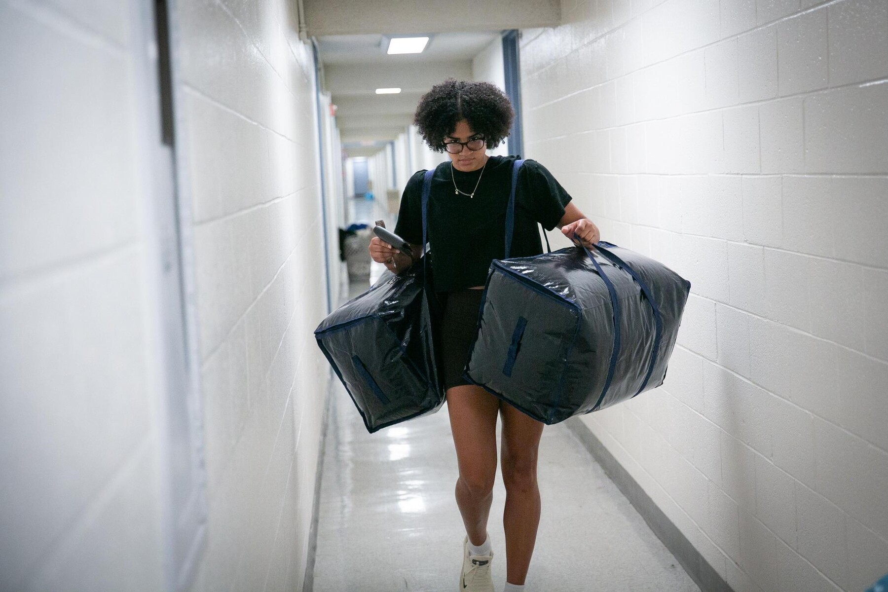 A student with two large bags walking down the hallway of their dorm. 