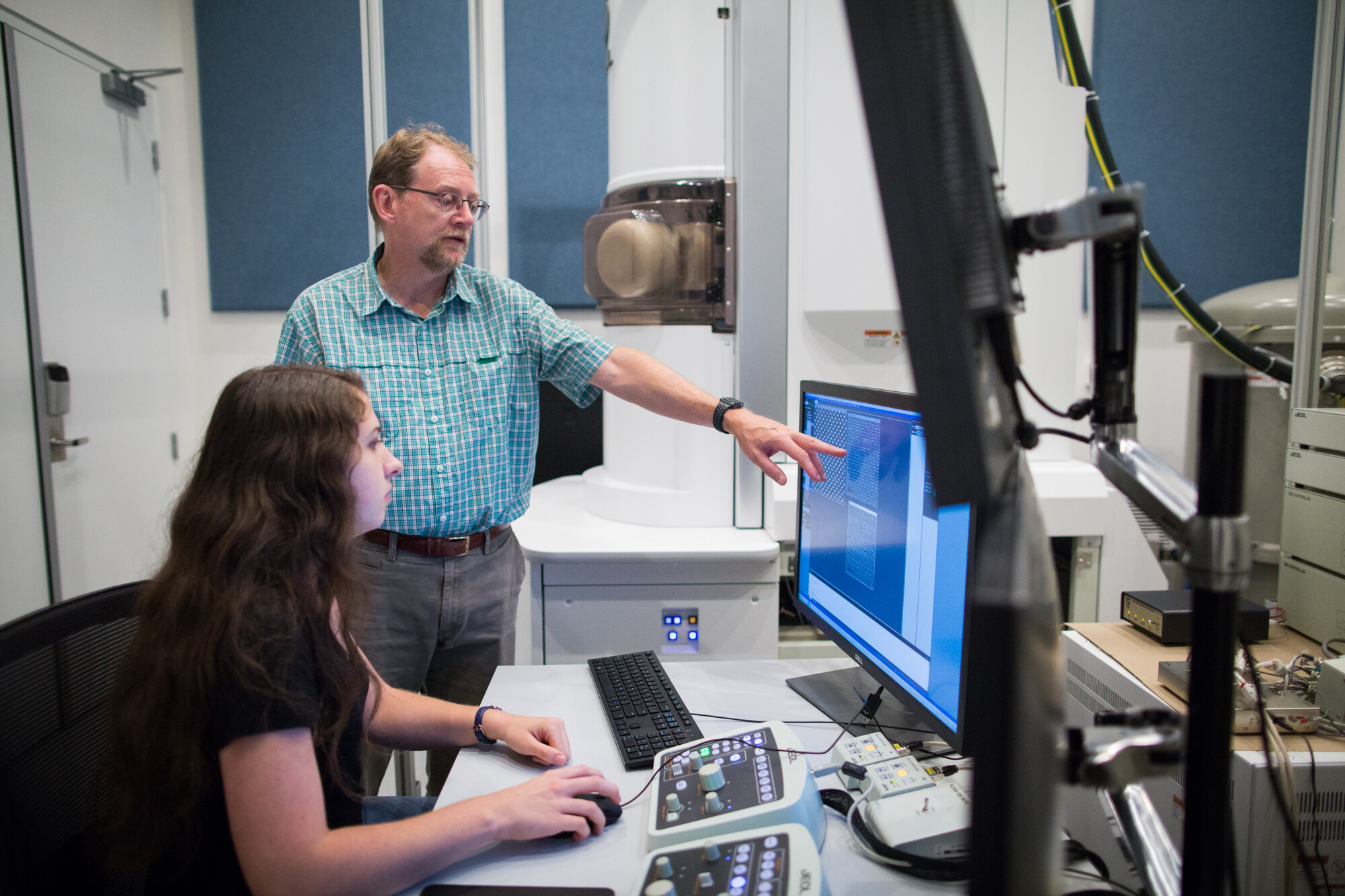 Two researchers look at a computer monitor, one person standing and the other seated, in a lab setting