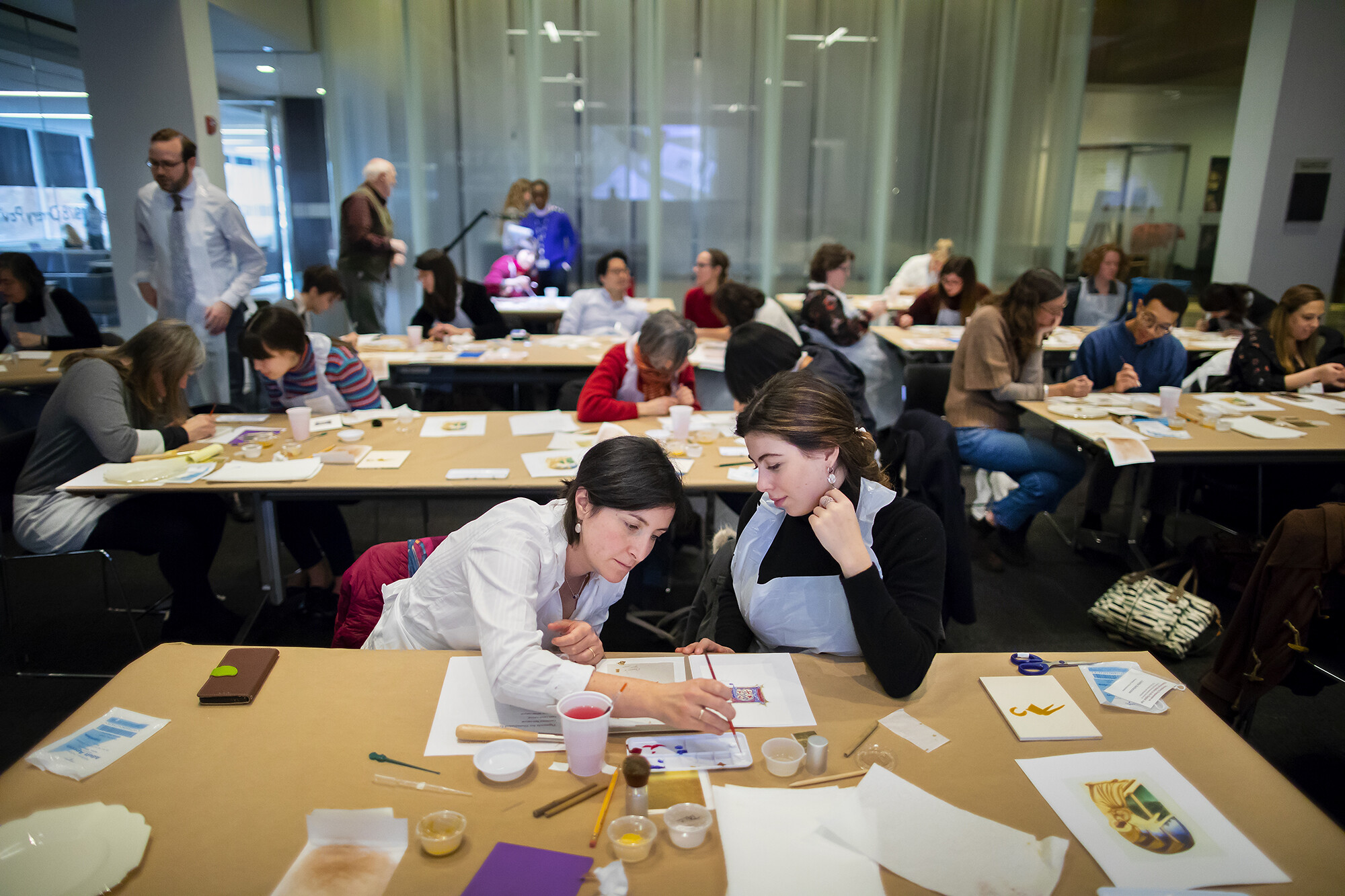 two women at a table working with art tools in the foreground with tables behind them and other students working on paper