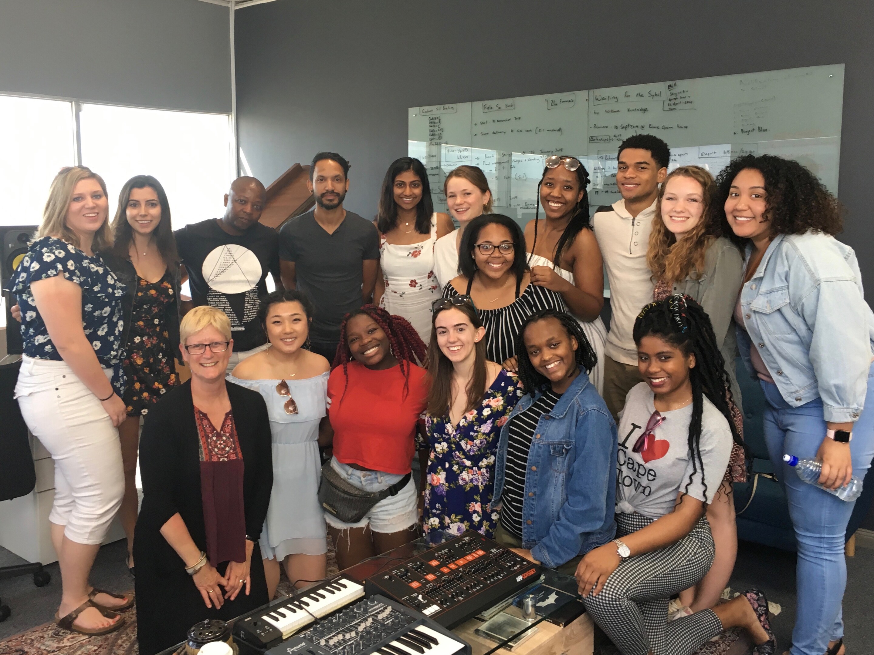 Group of students and professor gathered in studio of music composer with piano keyboard in front of them and a grand piano behind them. 
