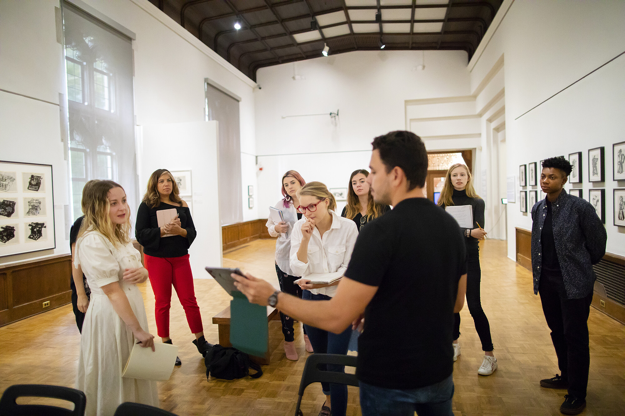 The artist is showing a student an iPad while other students look on, standing in a semicircle, in the Arthur Ross Gallery. 