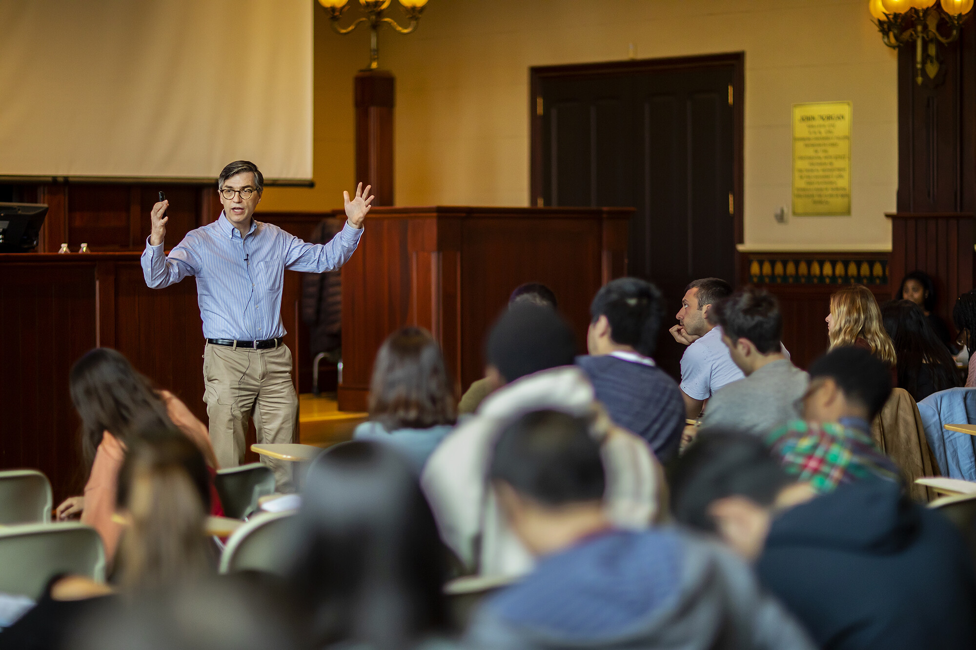 A man in a blue shirt and khakis gesturing in front of students sitting at desks.