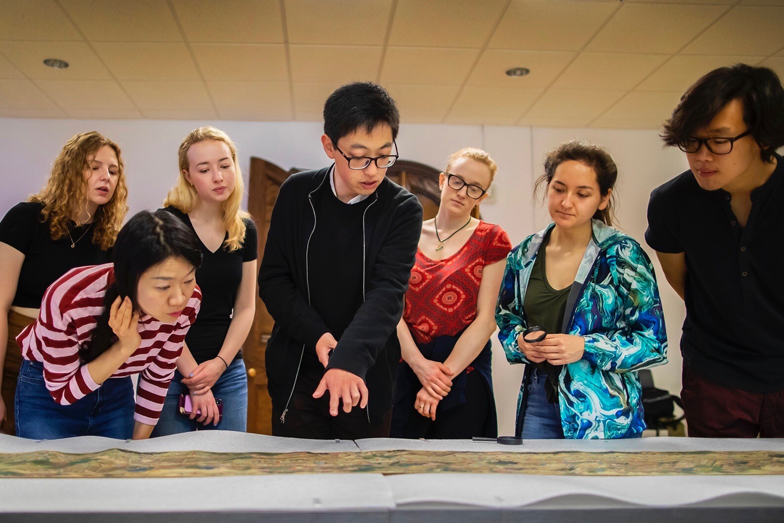 Seven students gathered together looking at a painted Chinese scroll unrolled on a table. 