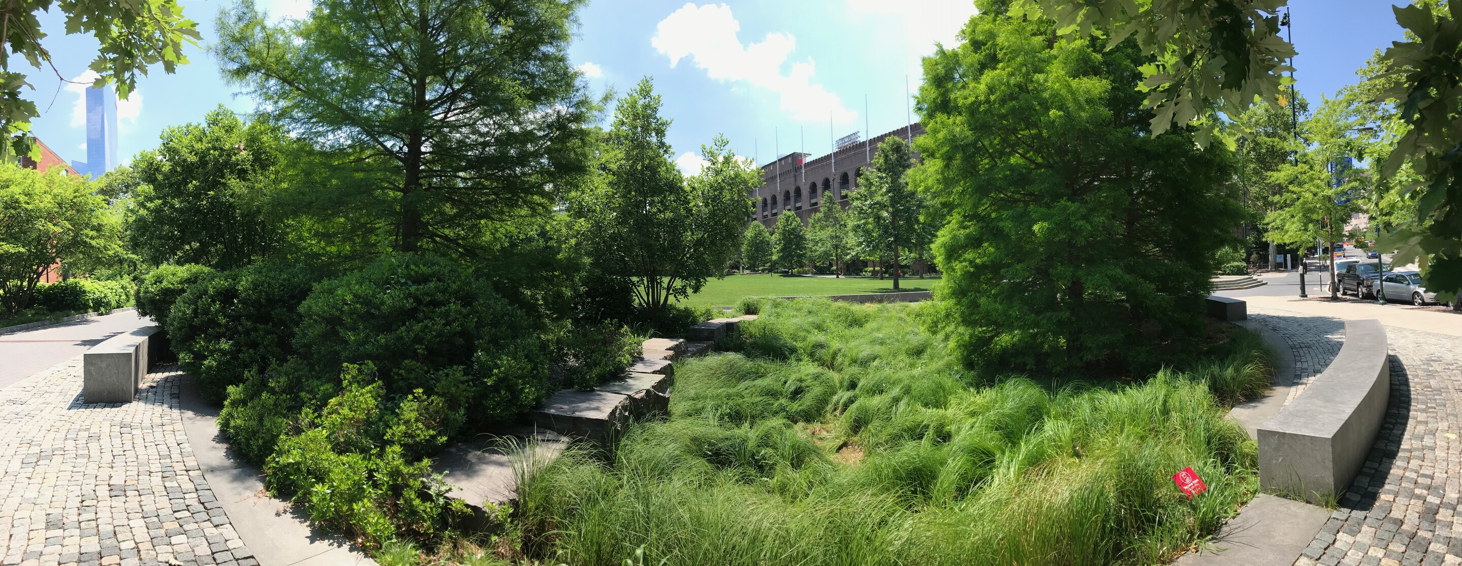 Landscape with large trees, grasses, a low stone wall and a brick stadium in the background
