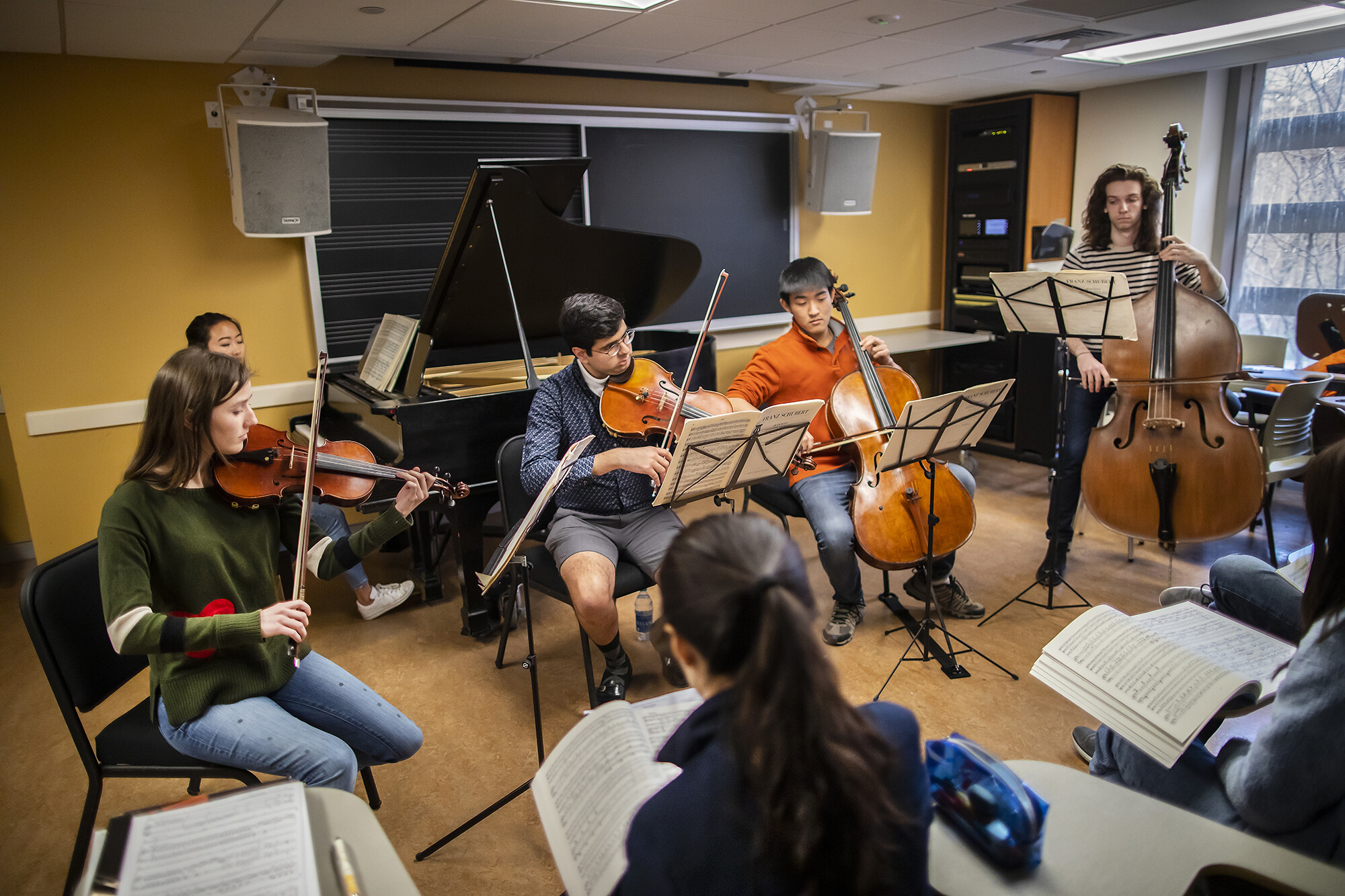Students play piano, violin, viola, cello, and bass, in front of music class.