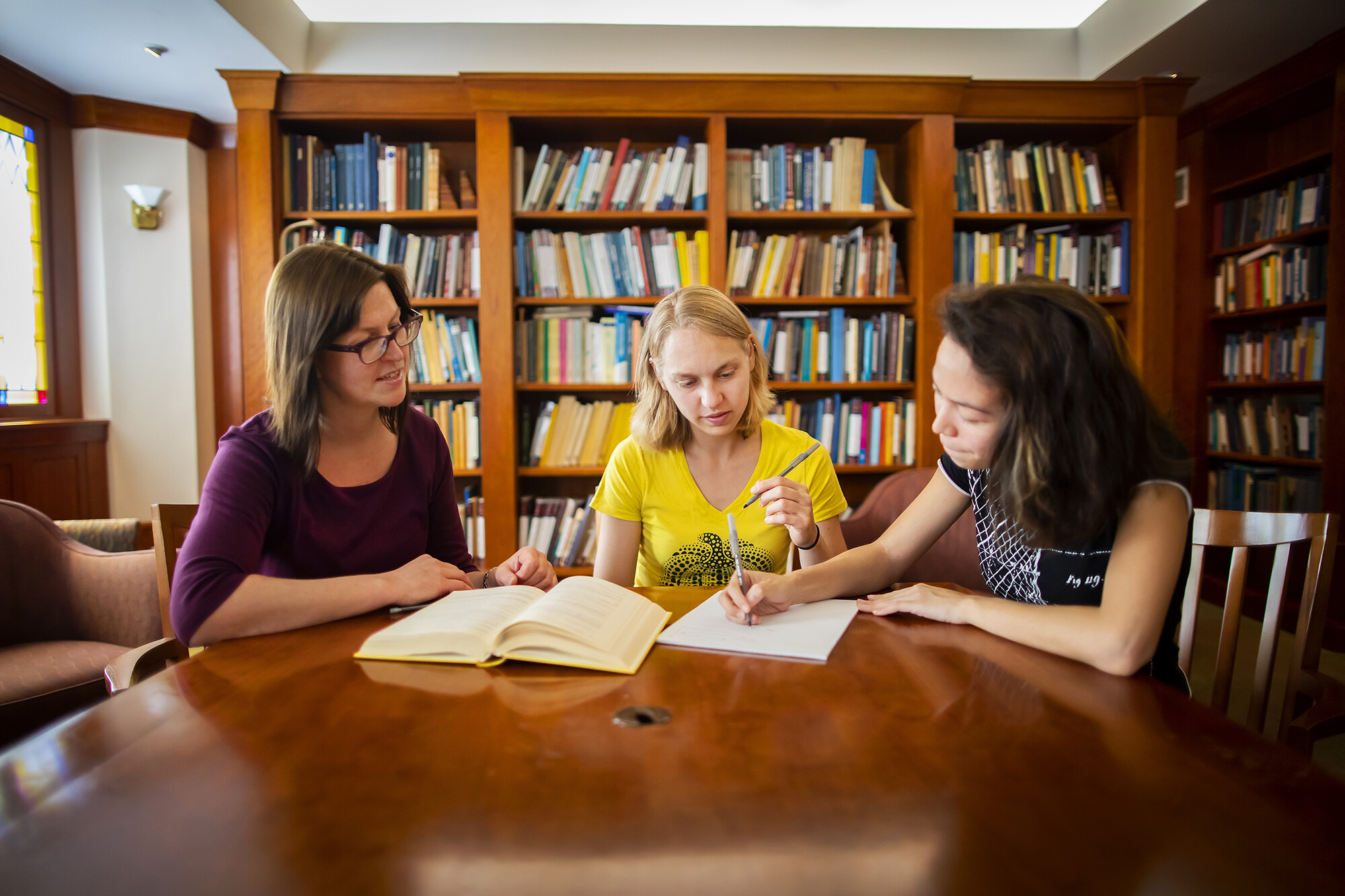three people sitting t a table inside a library writing with a pen and paper