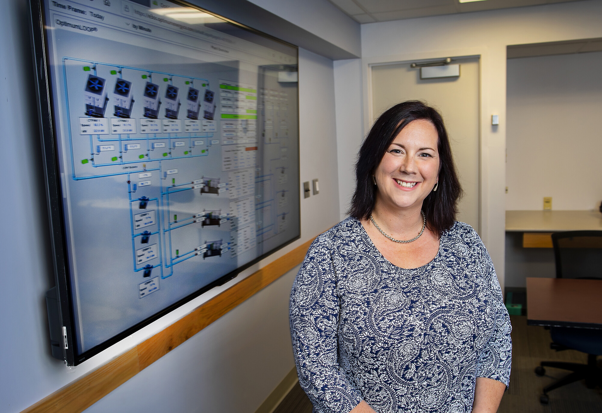 A smiling woman stands next to a screen that displays energy usage information for chiller plant system.