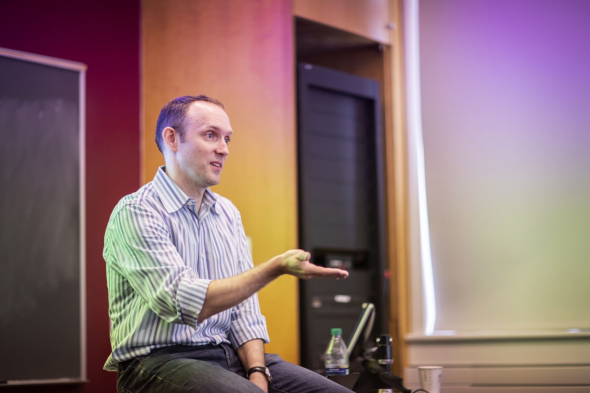 A seated lecturer in a classroom gestures and speaks to an unseen audience