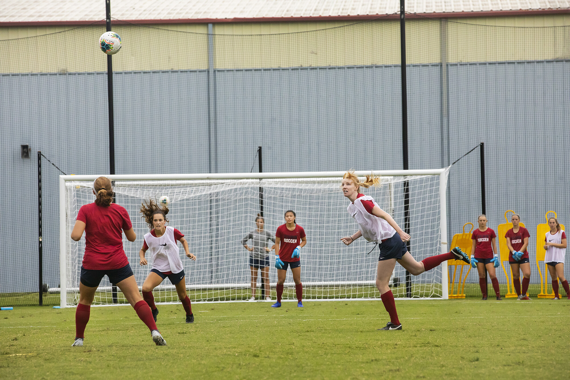Players practice on the field. The red team attempts to score on the white team. The ball is in the air.