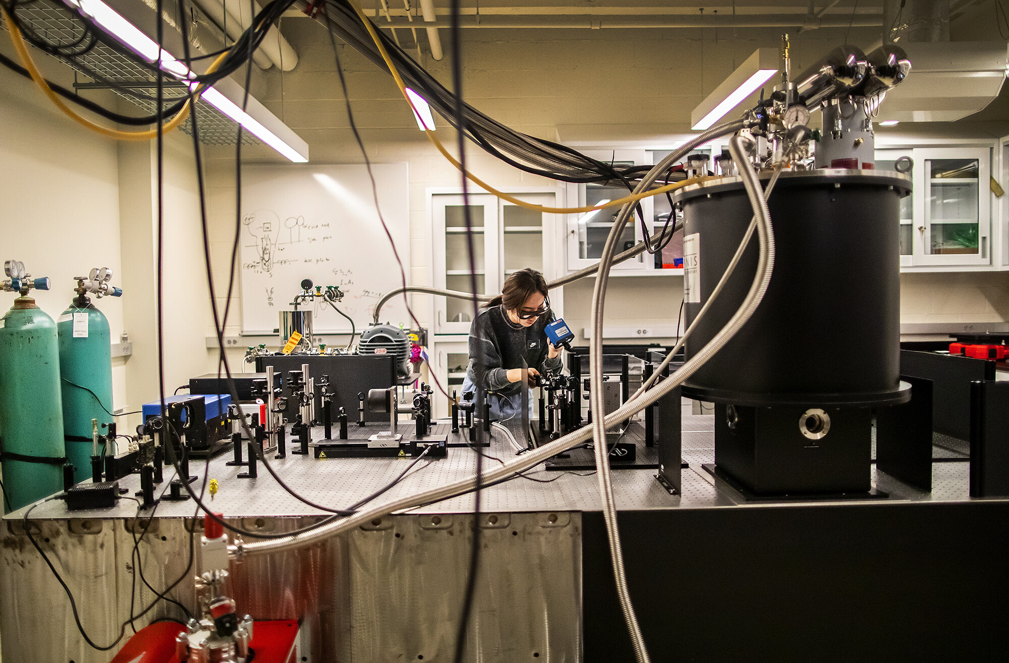 han looks through a scope at equipment on a large black optics table, surrounded by tubing, wires, an air pump, and gas canisters