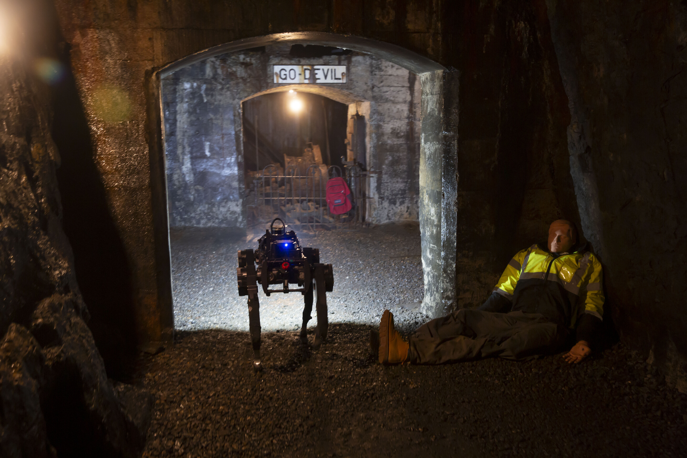 a legged robot walks down a mine past a dummy wearing a yellow vest and a red backpack that is hanging from a metal fence nearby