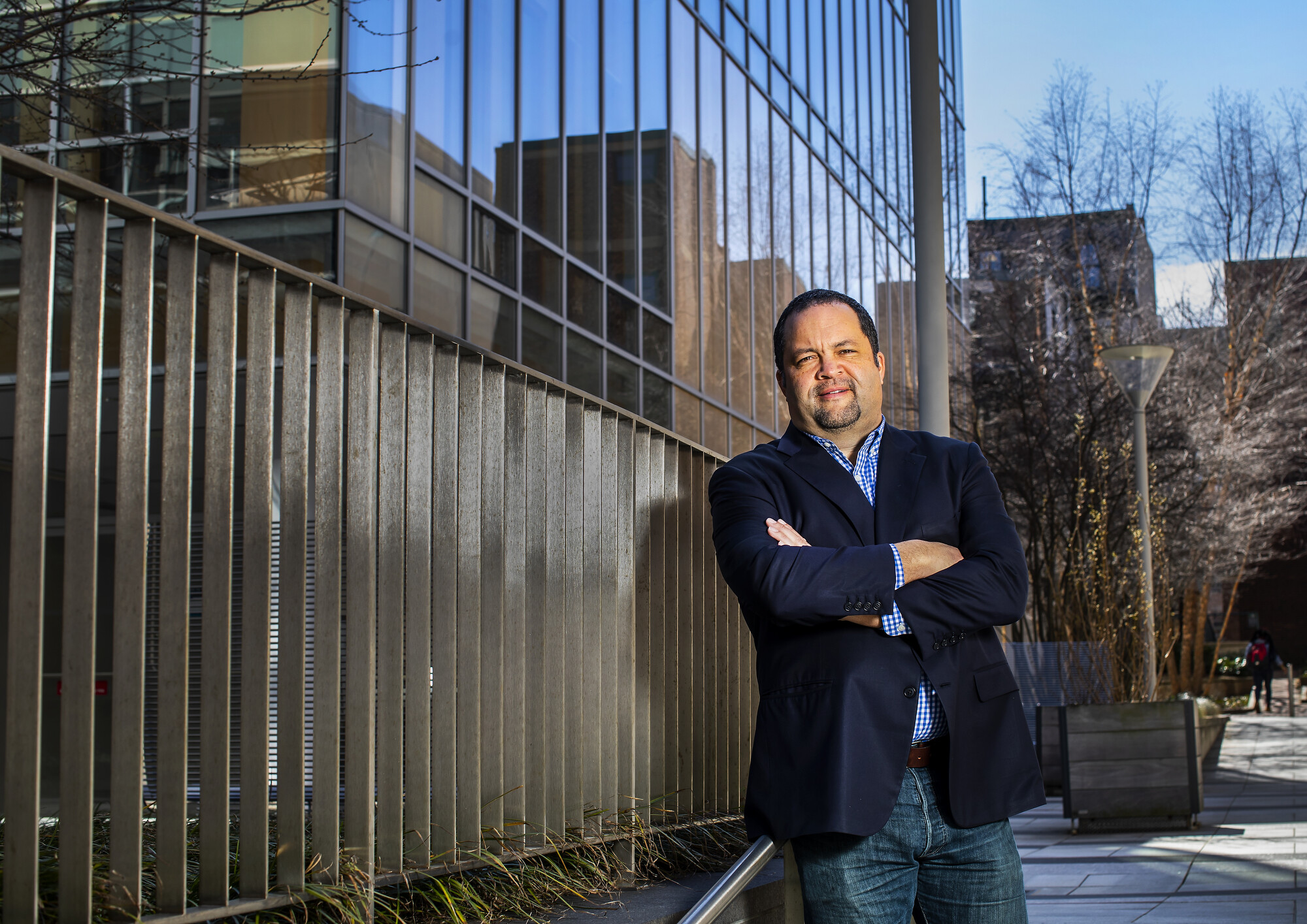 A person standing with arms crossed, in a courtyard next to a metal railing aside glass windows.