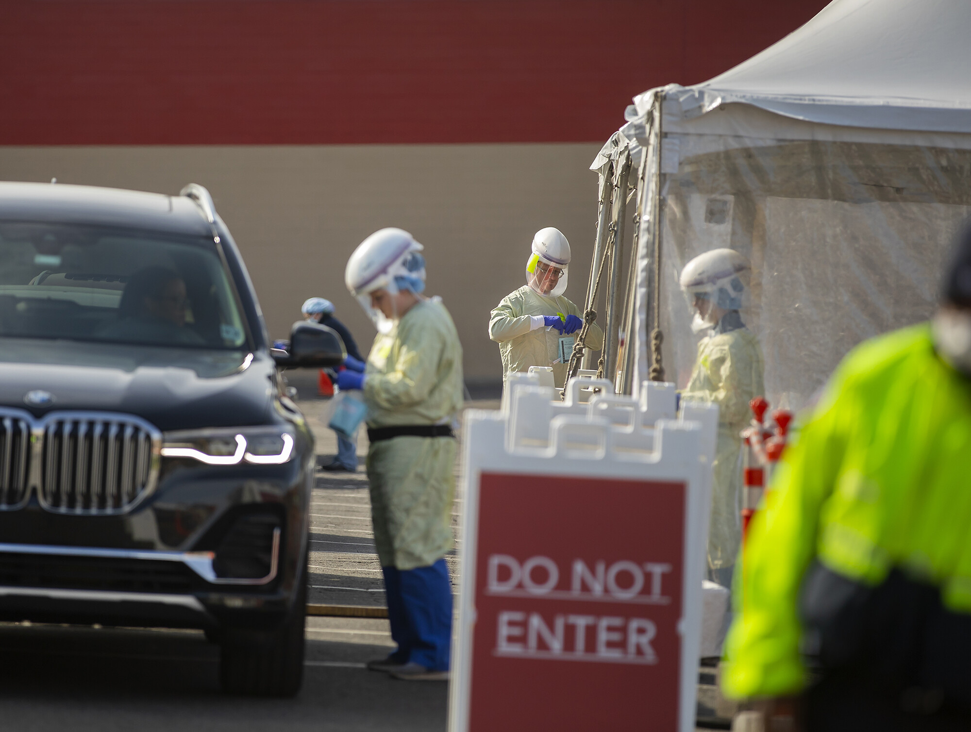 Three medical personnel outside in protective gear working at a drive-through COVID-19 testing site, a Don Not Enter sign in front of the vehicle