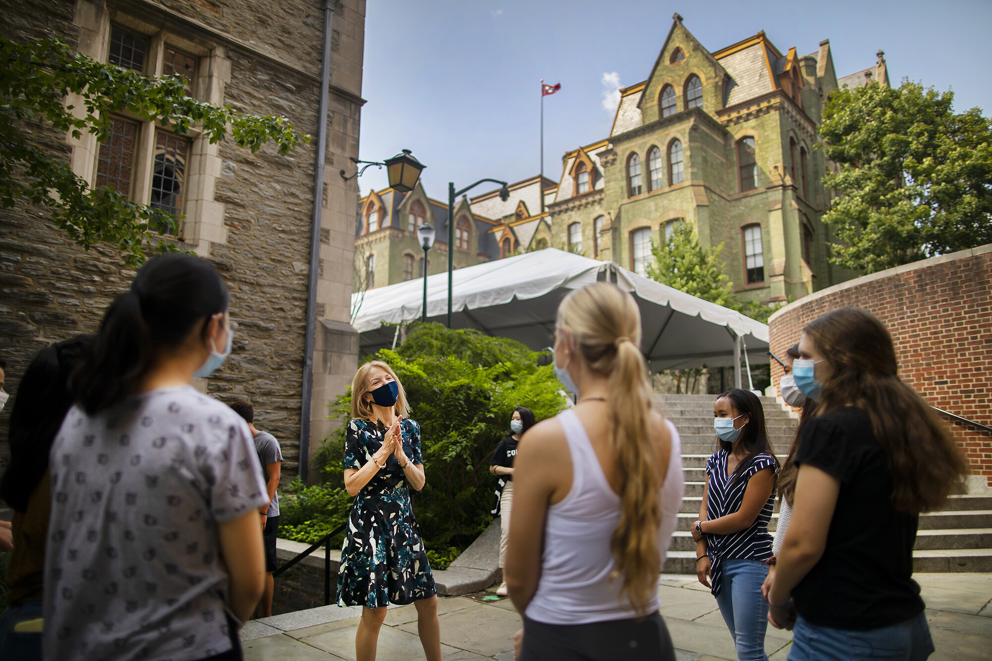 Penn President Amy Gutmann stands wearing a face covering in front of Houston Hall addressing four students wearing masks.