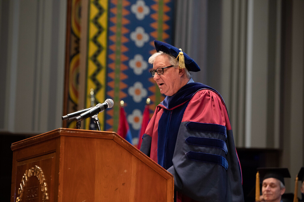 Steve Fluharty in full commencement regalia speaking at a podium.