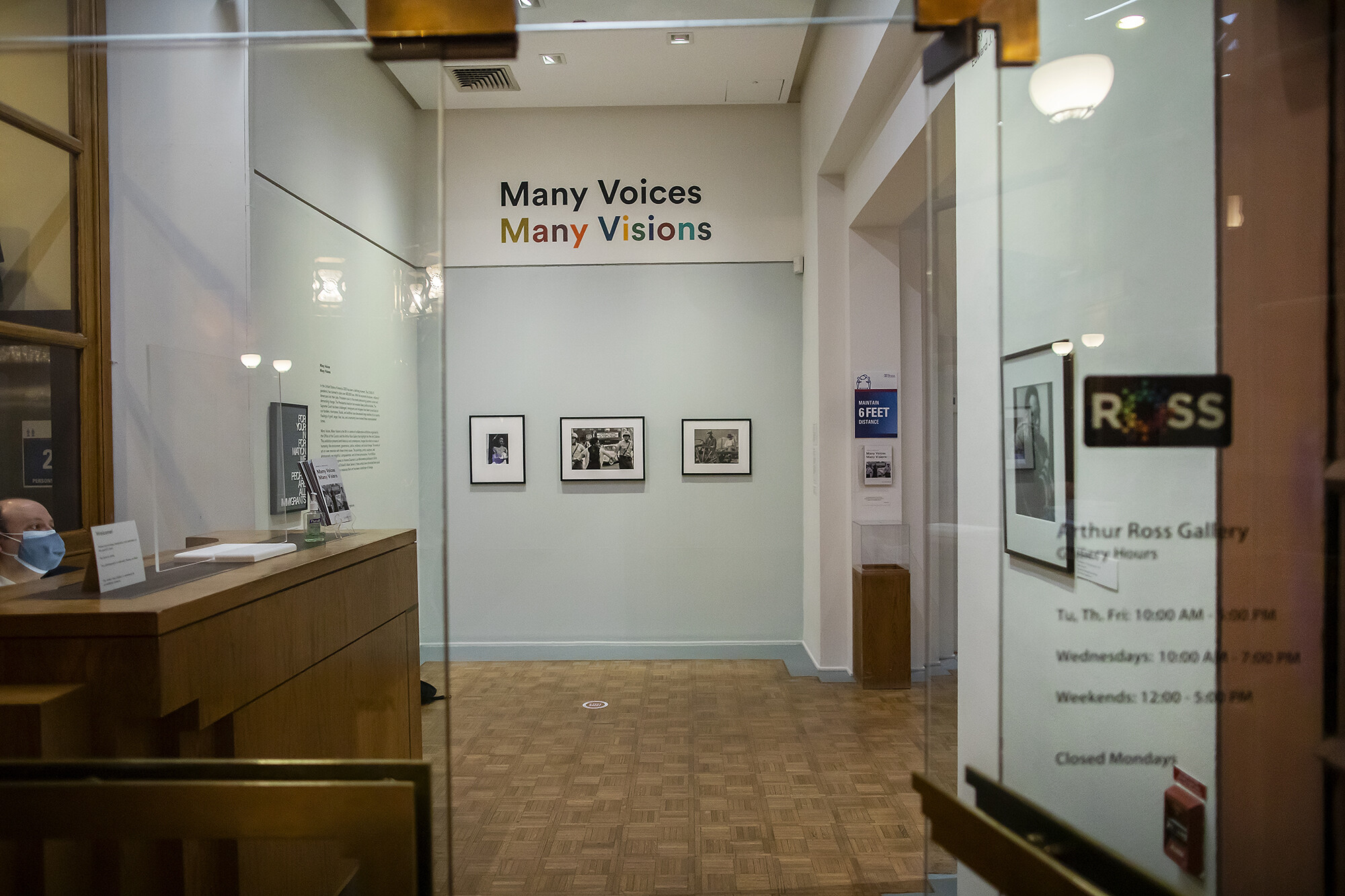 Glass doorway with guard seated at desk to left with three photographs on a wall with the words Many Voices Many Visions above.